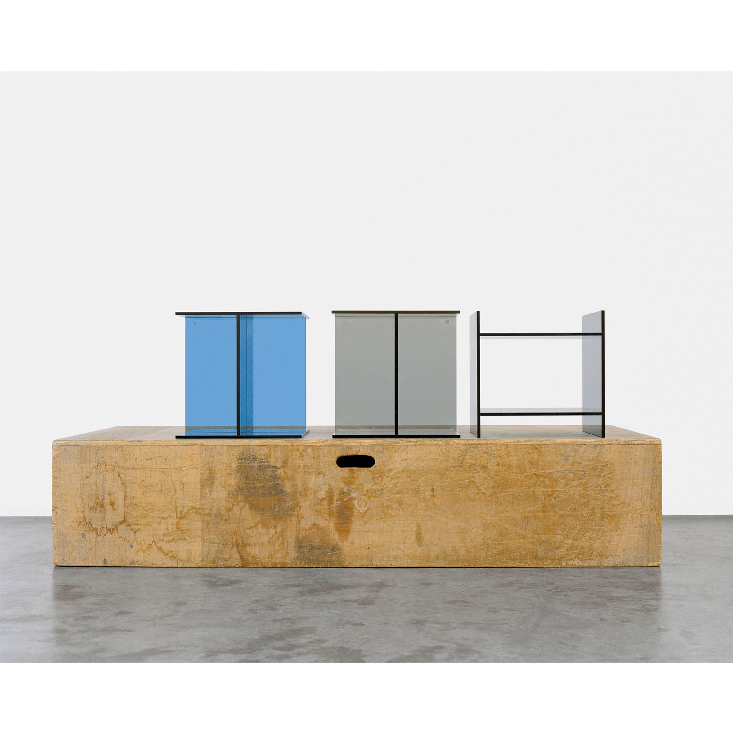 The E15 CT08 VIER features three minimalist, box-shaped sculptures crafted from solid colored glass in blue, gray, and clear hues, elegantly displayed on a wooden platform set against a plain white backdrop.