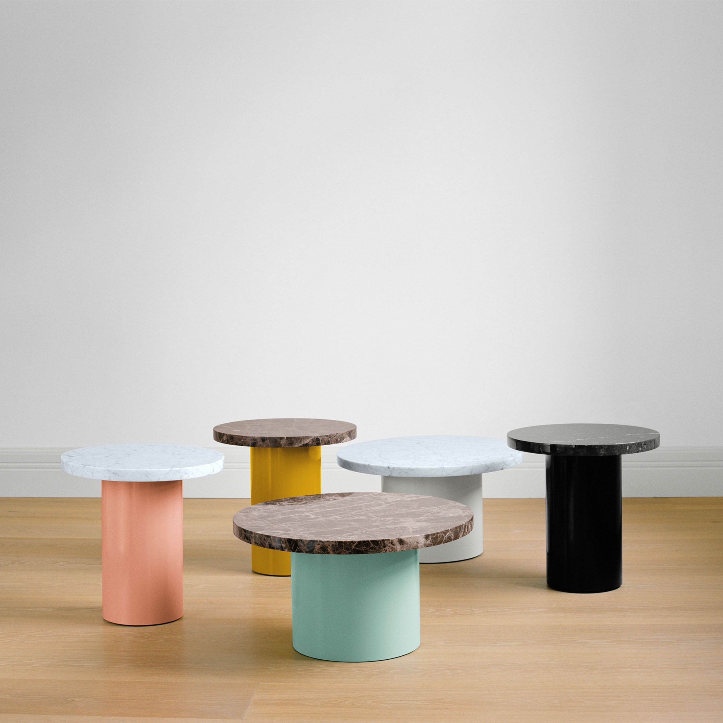 The CT09 ENOKI collection by E15 consists of five tables with vibrant cylindrical bases in peach, green, yellow, black, and white topped with rich marble. These modern pieces are set on a solid wood floor against a plain light gray wall.