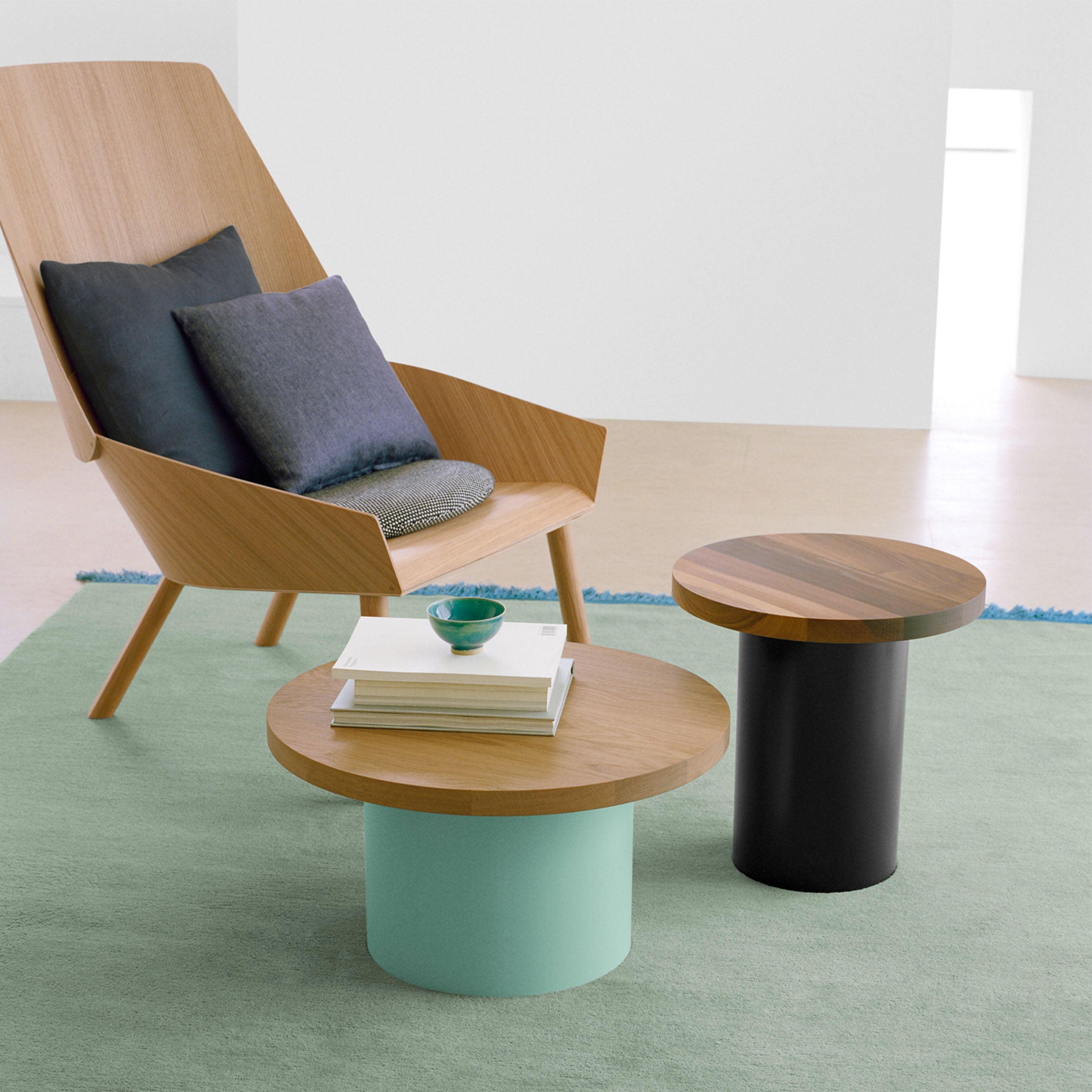 In a modern living room, a wooden armchair with gray and blue cushions is complemented by two E15 CT09 ENOKI tables, one light green and the other black, both featuring cylinder bases. These sit on a lush green carpet, adorned with a small bowl and books for added serenity.