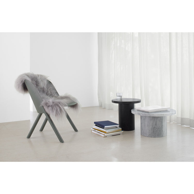 In a minimalist room, a gray chair with a furry throw sits next to two CT09 ENOKI tables by E15. The rich marble on the black surfaces adds elegance, complemented by book stacks. A water glass rests on one table, and sheer curtains softly cover the large window behind.