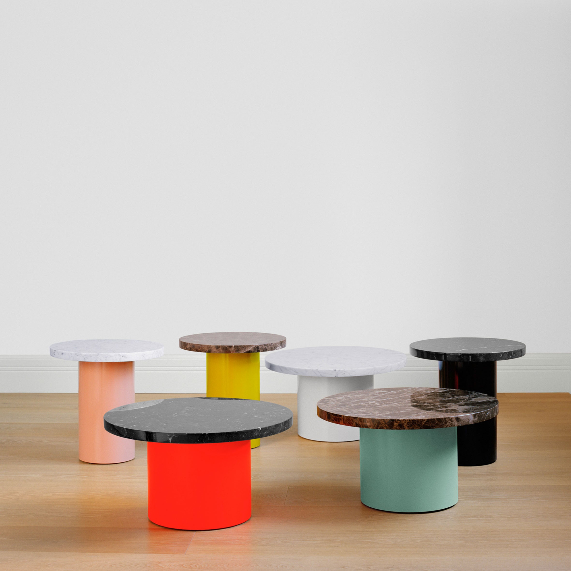 The CT09 ENOKI collection by E15 features six tables with vibrant cylindrical bases in orange, yellow, white, black, and green paired with luxurious marble tops. They are elegantly showcased in a minimalist room with solid wood flooring and pristine white walls.