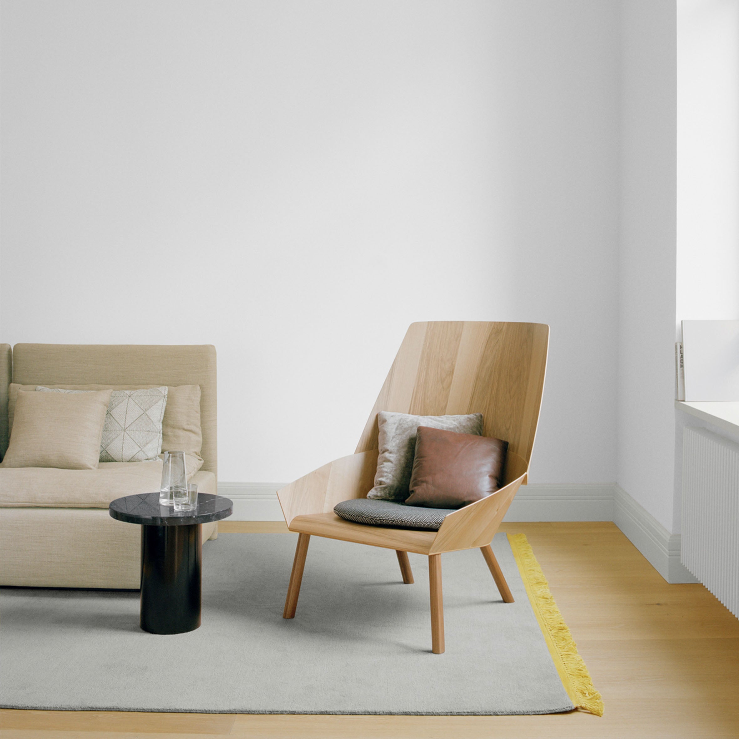 A minimalist living room with a beige sofa, wooden chair, and a small CT09 ENOKI by E15 on a gray rug with yellow trim. The solid wood table holds a glass and vase. Cushions adorn the seating, while natural light floods in through a large window.
