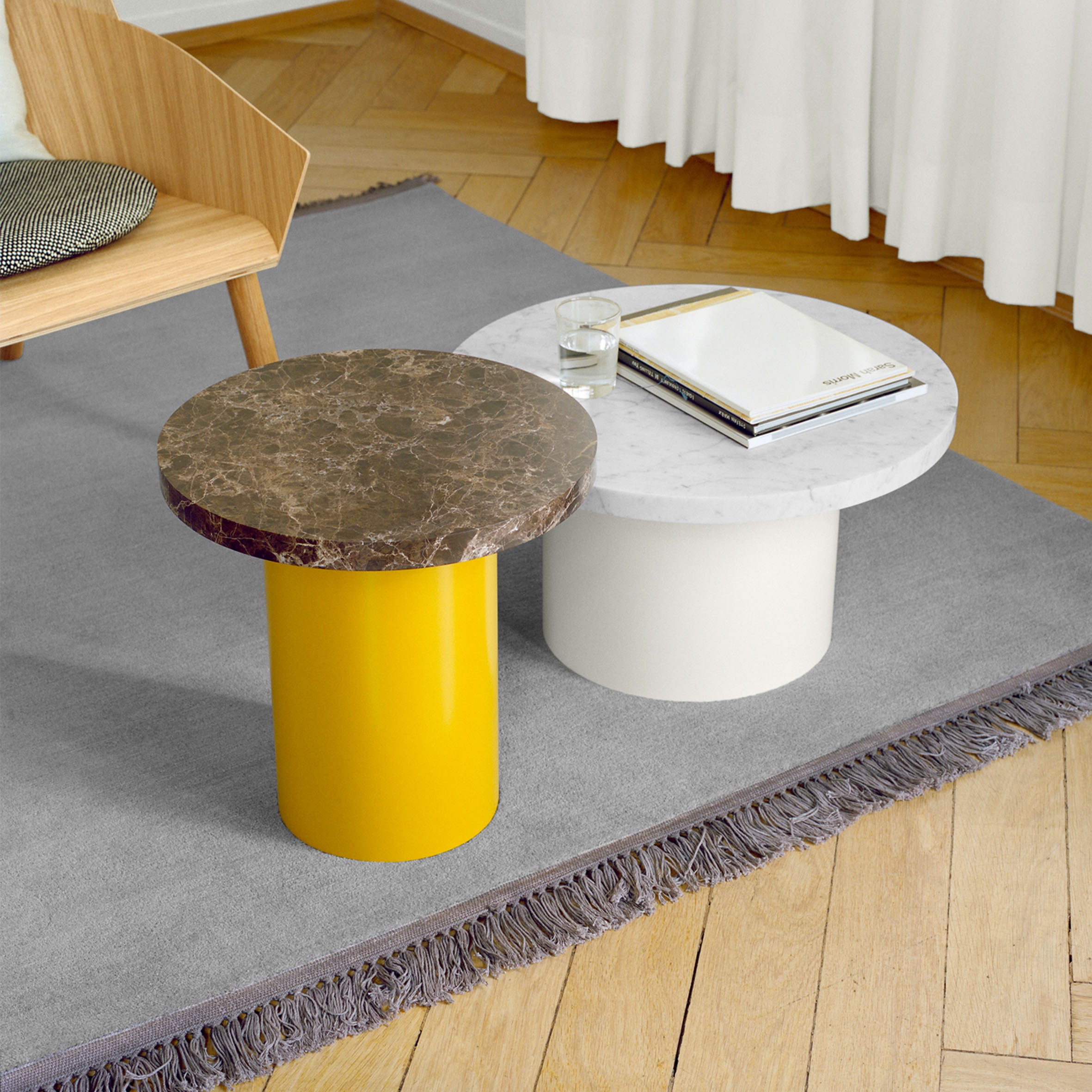 A modern living room with a wooden floor and light gray rug showcases two E15 CT09 ENOKI tables: one with a yellow base and rich marble top, the other featuring a white base adorned with books and a glass of water on its pristine white marble top. A solid wood chair is nearby.