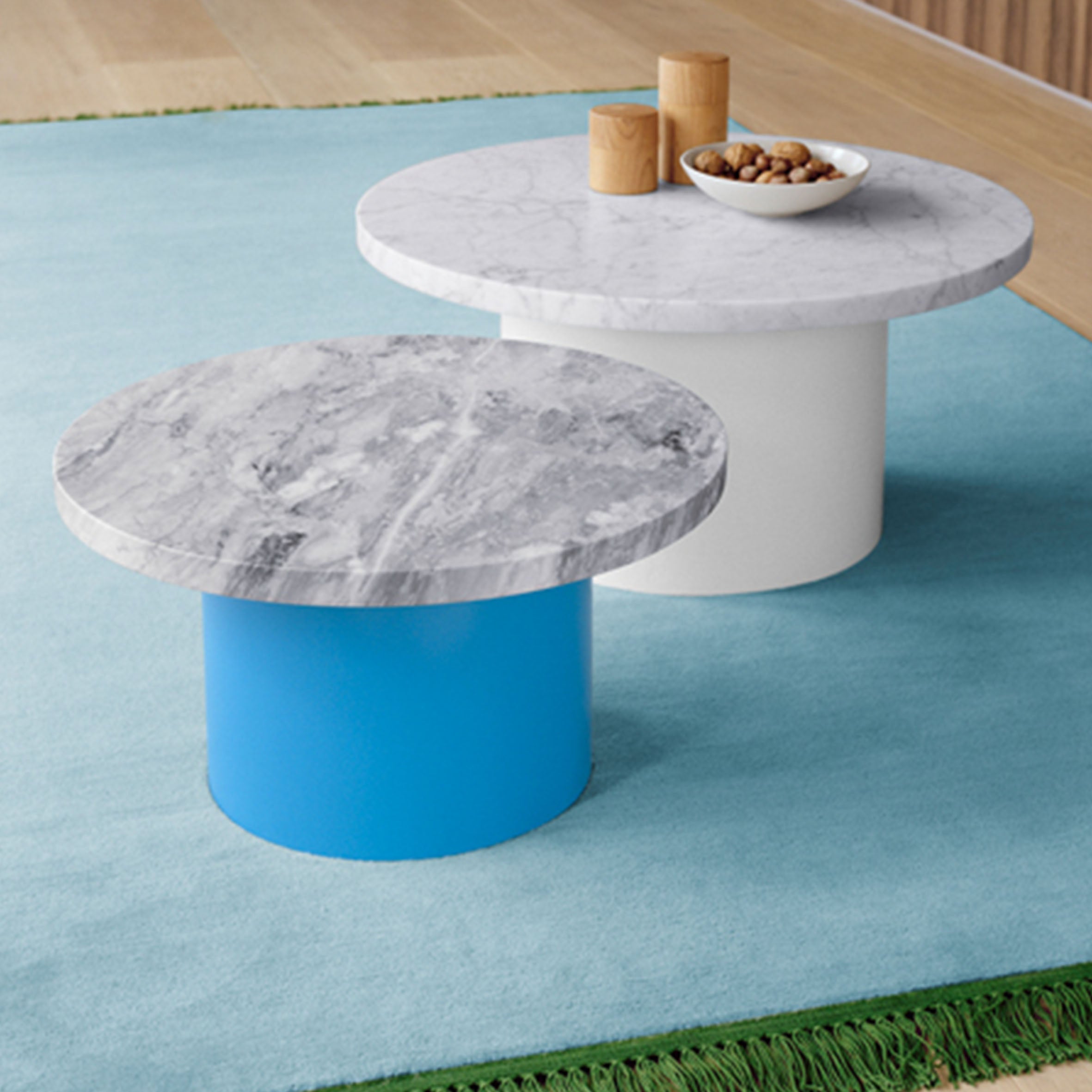 Two CT09 ENOKI tables from E15, featuring rich marble tops—one with a white base and another with a blue base. The taller table holds three cylindrical wood containers and a nut bowl, all placed on a blue-green carpet over wood flooring.