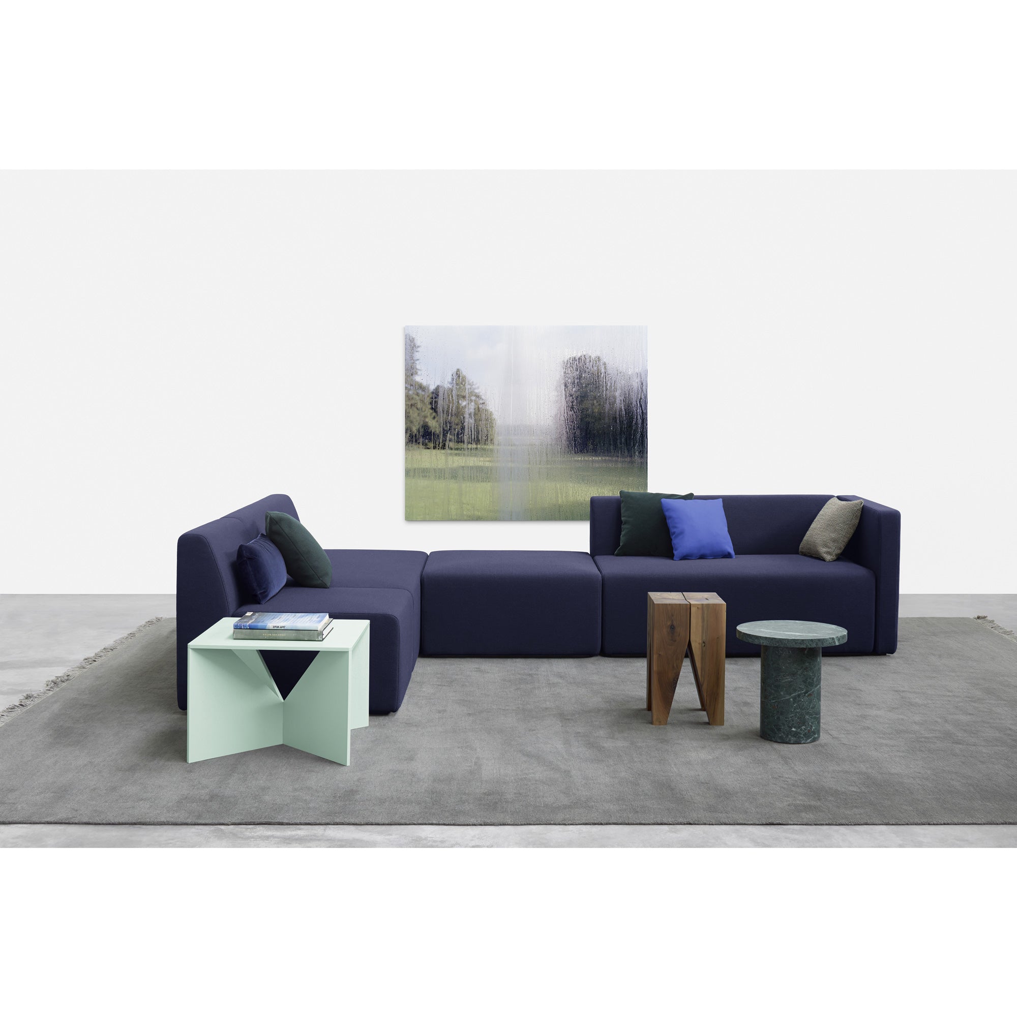 In a modern living room, a navy-blue sectional sofa with cushions is paired with a geometric green coffee table from E15s SELECTED line. A wooden stool and the E15 ENOKI MIGOTO round marble side table sit on a gray rug, under a landscape canvas on the white wall.