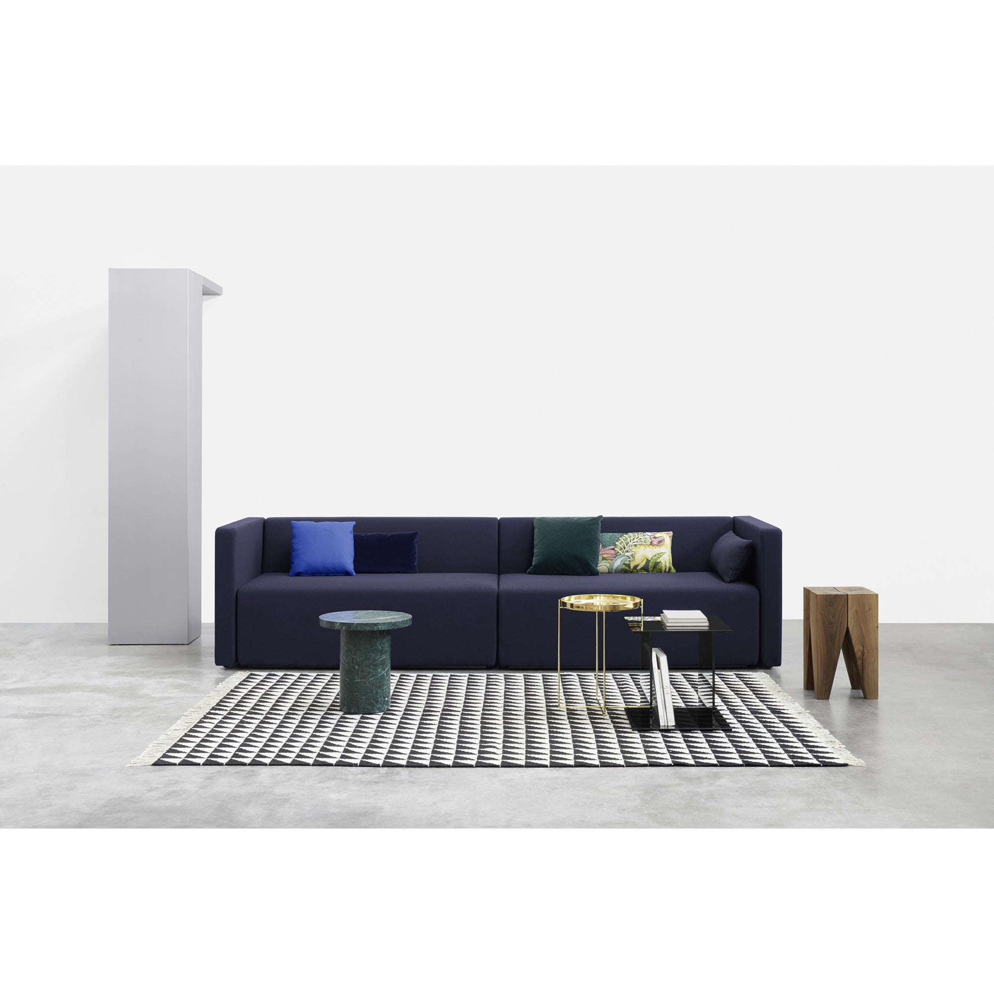 A modern living room showcases a dark blue sofa with colorful cushions, a black and white patterned rug matching the ENOKI MIGOTO green marble coffee table by E15, and smaller wooden tables. The minimalist decor features concrete flooring and white walls, enhanced by an E15 SELECTED side table.