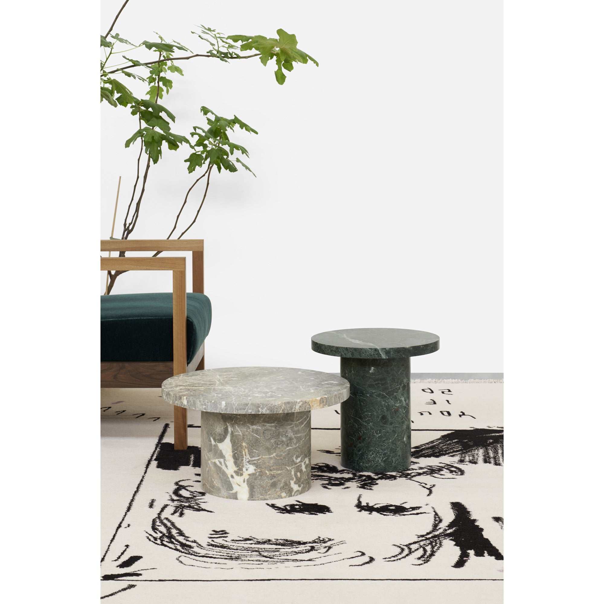 A gray and dark green round marble table sit elegantly on a white mat with abstract black designs, accompanied by a wooden chair with a green cushion and a leafy plant against a plain white wall, embodying the E15 brands ENOKI MIGOTO style.
