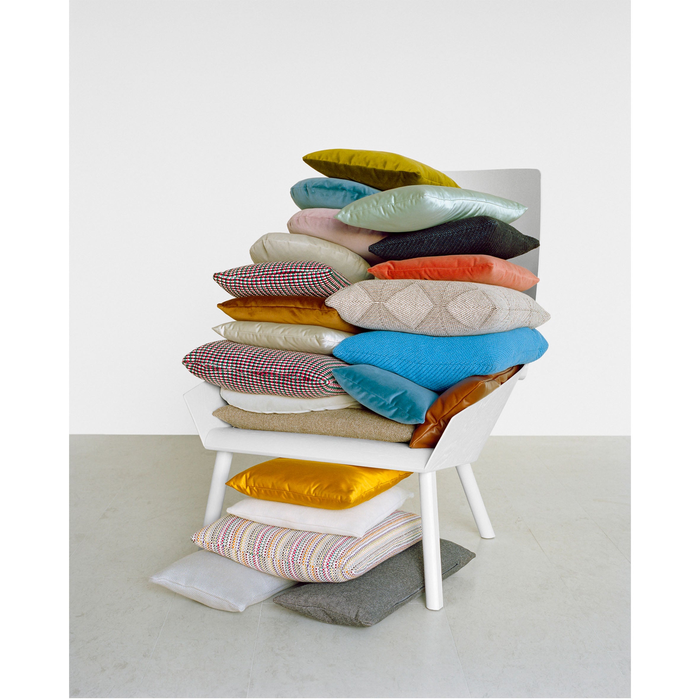 The CU06 NIMA by E15 is a white chair adorned with a variety of colorful accessory cushions, featuring houndstooth and diverse textures in fabric and leather. Some cushions are scattered on the floor, enhancing the vibrant contrast against a plain white background.