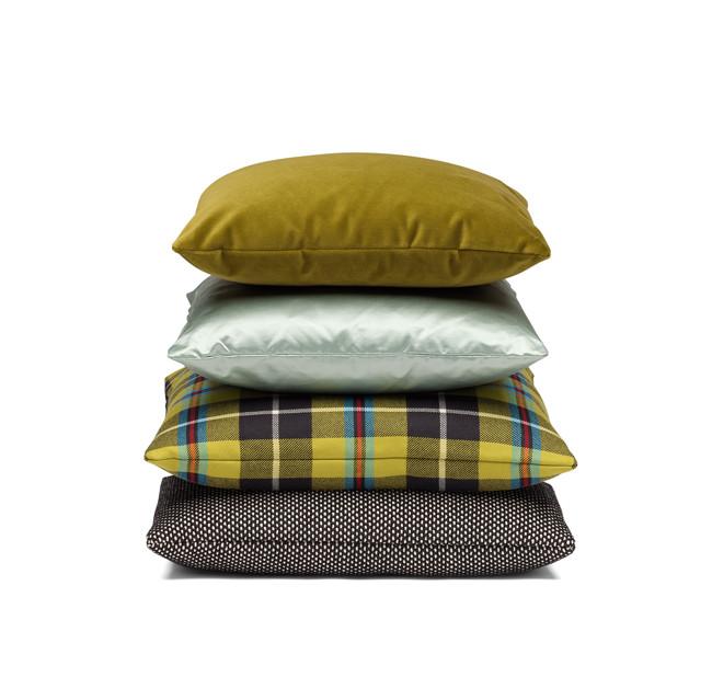 A stack of four E15 CU06 NIMA cushions includes a plain olive green pillow on top, a light mint accessory cushion beneath, yellow/blue/burgundy plaid next, and a black pillow with white polka dots at the bottom.