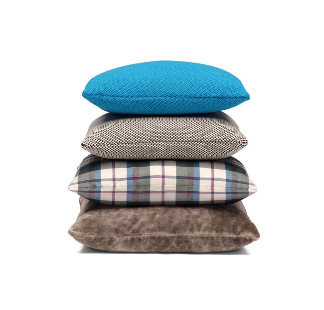 A vertical stack of four E15 CU06 NIMA pillows displays diverse seating elements: a blue top, beige with small dots second, blue and beige plaid pattern third, and a bottom in textured brown fabric—all set against a white background.