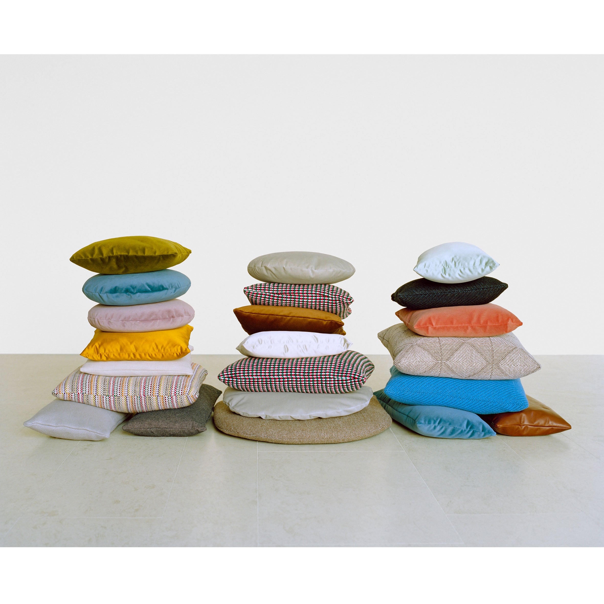 Three stacks of CU06 NIMA pillows by E15 in various sizes and patterns are displayed on a light floor against a white background. With vibrant yellow, blue, beige, and patterned designs, these accessory cushions create a visually diverse arrangement.