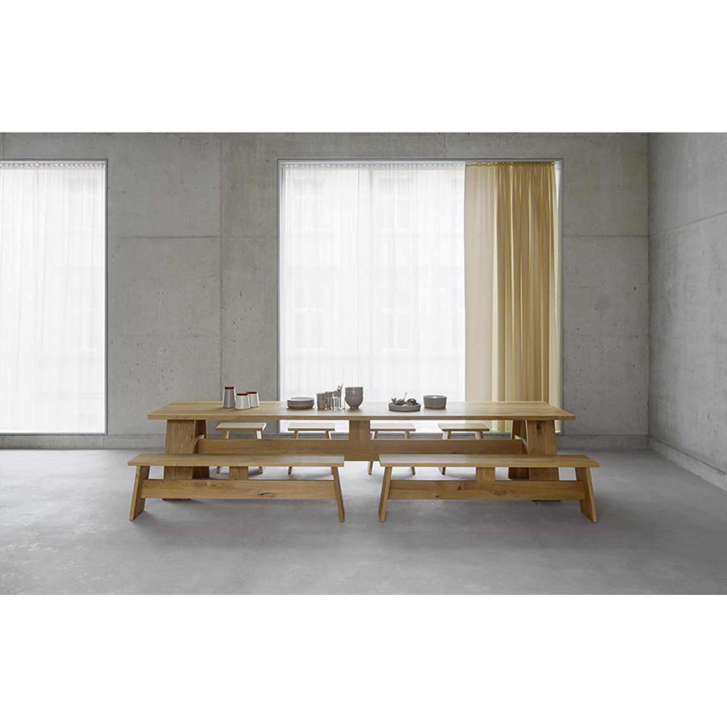 A minimalist dining area highlights an elegant E15 DC01 FAYLAND solid wood table and benches. Large windows with sheer white curtains accent the cement wall, while simple settings of cups, plates, and books amplify the airy ambiance inspired by David Chipperfields refined design.