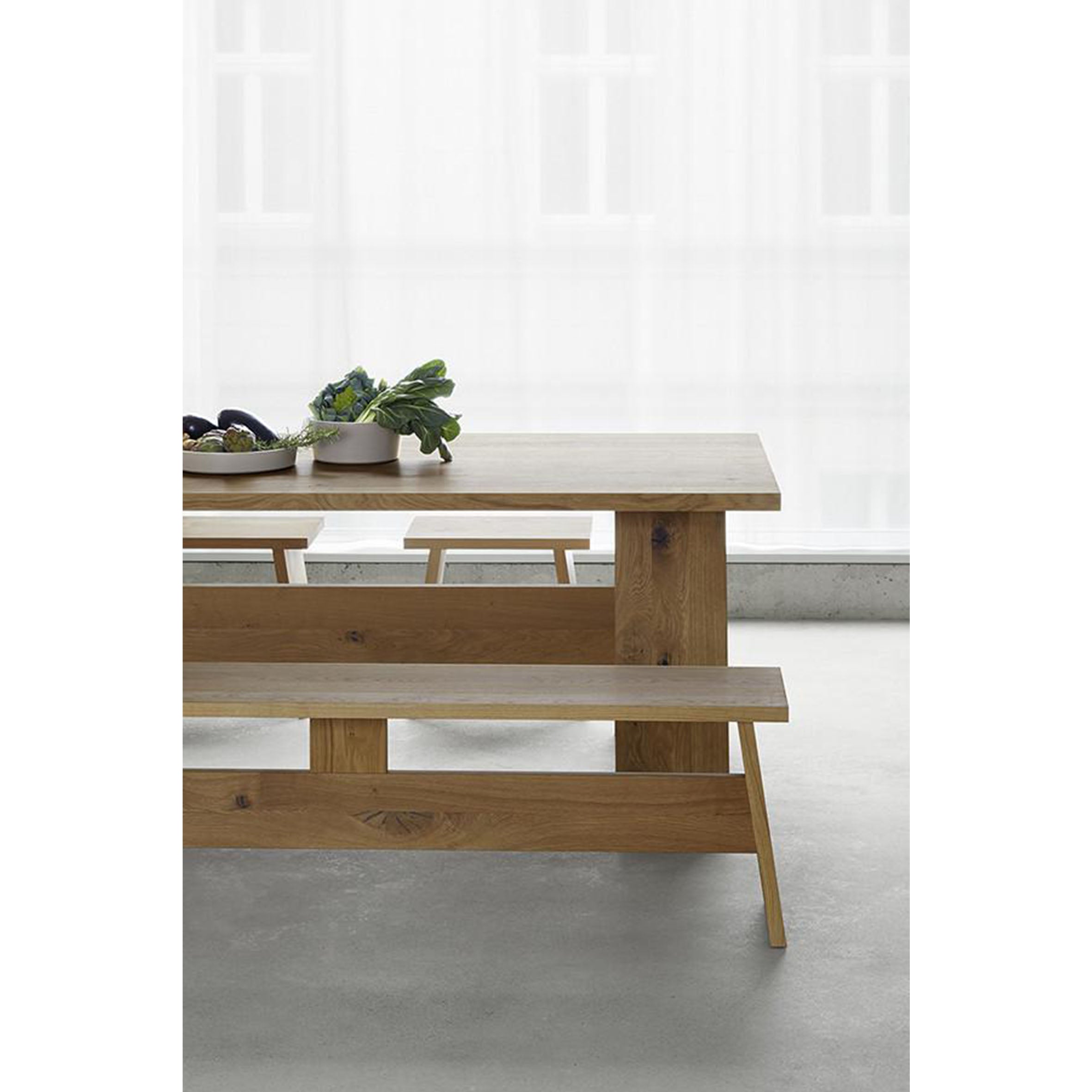 The DC02 FAWLEY by E15, a wooden dining table made from European walnut, pairs with a solid wood bench in a minimalist room. It stands elegantly by a large window with sheer curtains. Bowls of leafy greens on the table add vibrant contrast to the settings sleek black silhouette.