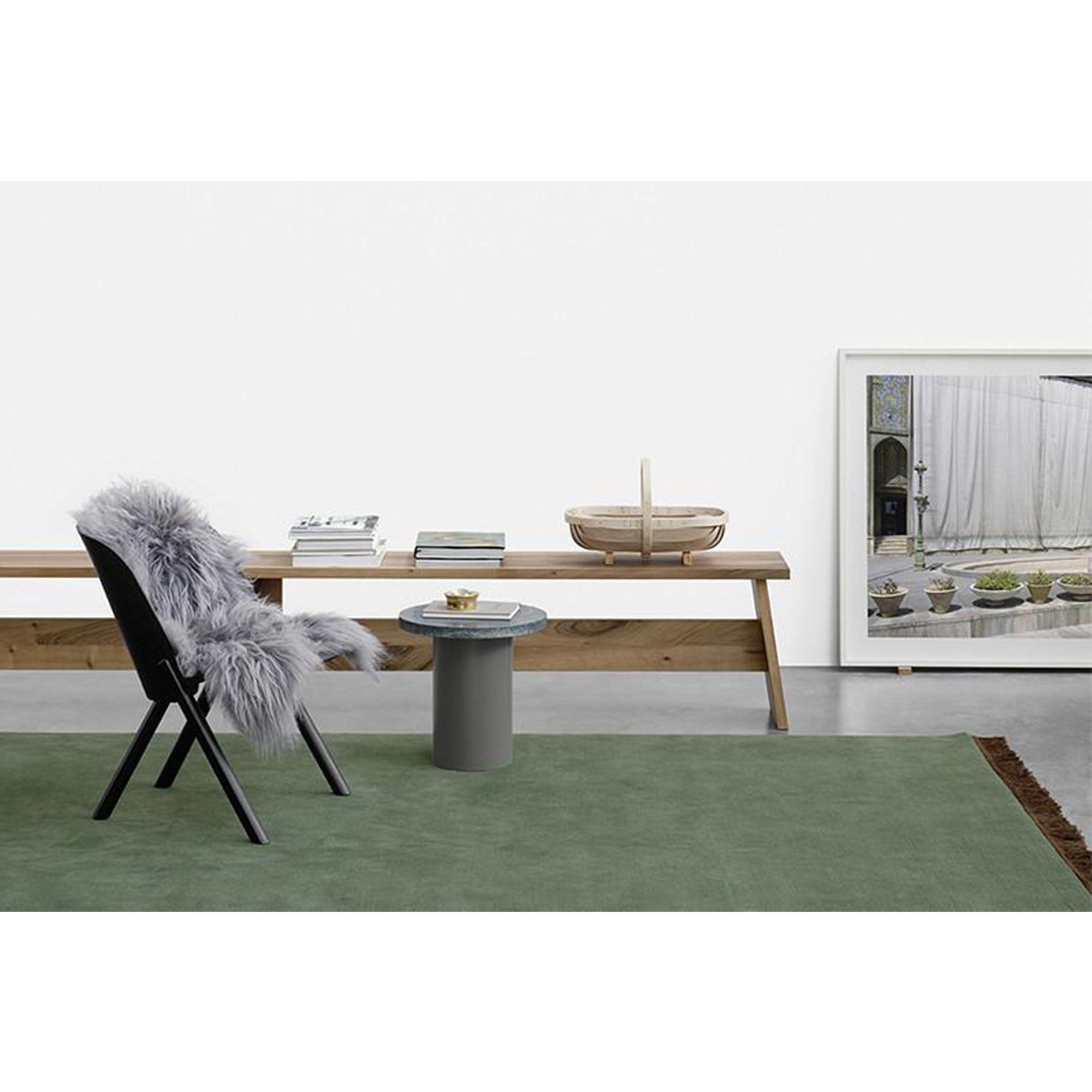 A minimalist interior features a black chair with a gray throw, a small round table, and an E15 solid wood DC02 FAWLEY bench adorned with books. Nearby is a wicker basket, and framed art leans against the white wall on a green rug inspired by David Chipperfields design sensibilities.