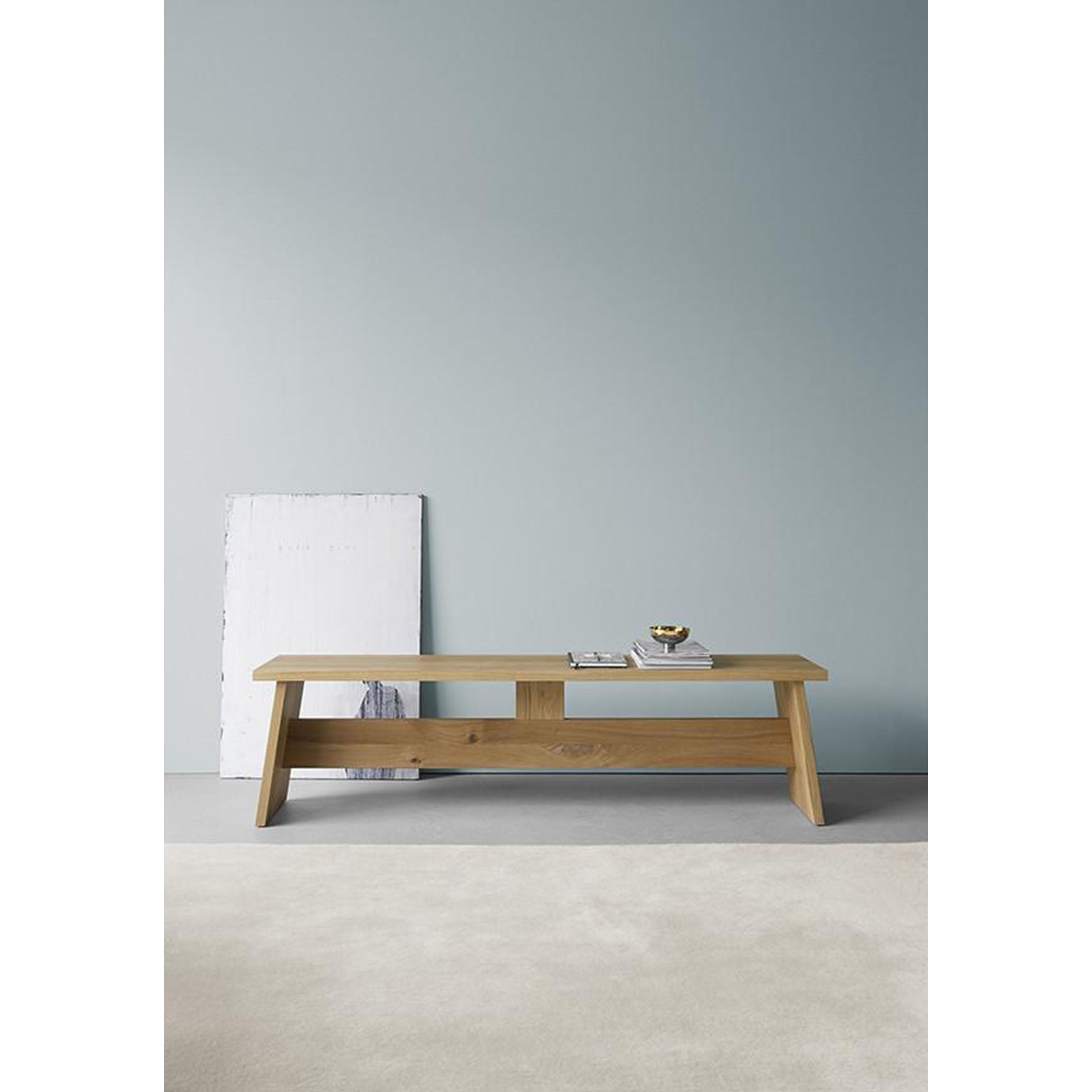 The DC02 FAWLEY, a minimalist E15 solid wood bench with angled legs, is set against a light gray wall with a white, slightly worn board leaning behind it. Reminiscent of David Chipperfields style, it supports a small bowl and several books on the light-colored carpeted floor.