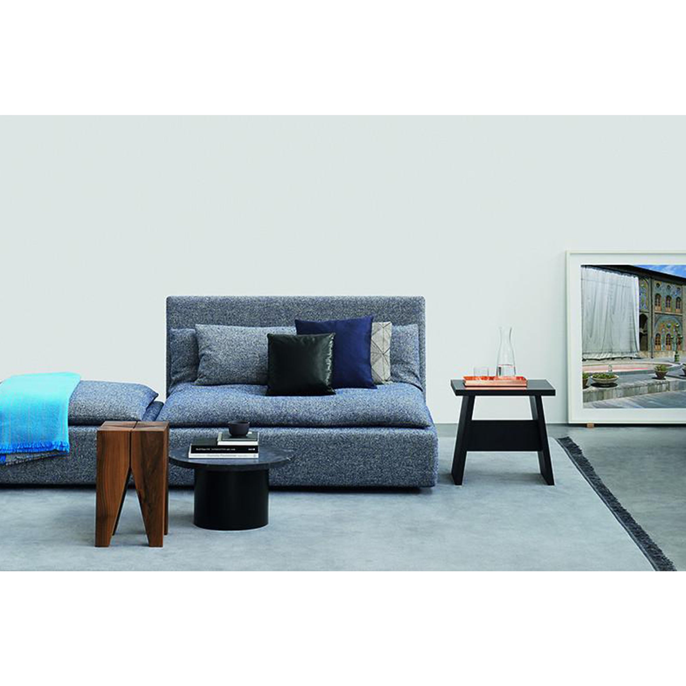 A modern living room showcases a blue sofa and chaise lounge, with two solid wood side tables holding books and decor. An art piece rests on the floor, accented by a blue throw and black, gray, and blue cushions, highlighted by the sleek presence of an E15 DC03 LANGLEY stool.