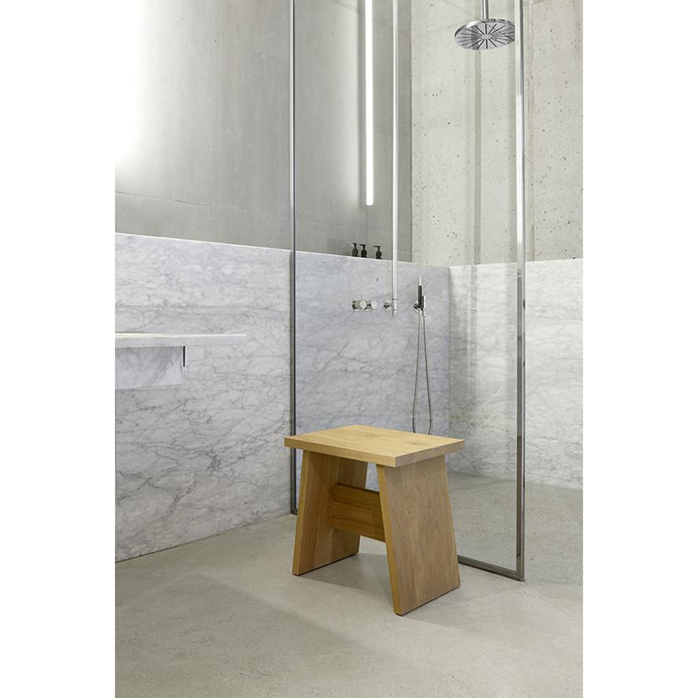 A minimalist bathroom showcases a walk-in shower with a glass partition and ceiling-mounted showerhead. An E15 DC03 LANGLEY stool, crafted from European walnut, rests on the concrete floor, while white marble tiles partially adorn the walls.