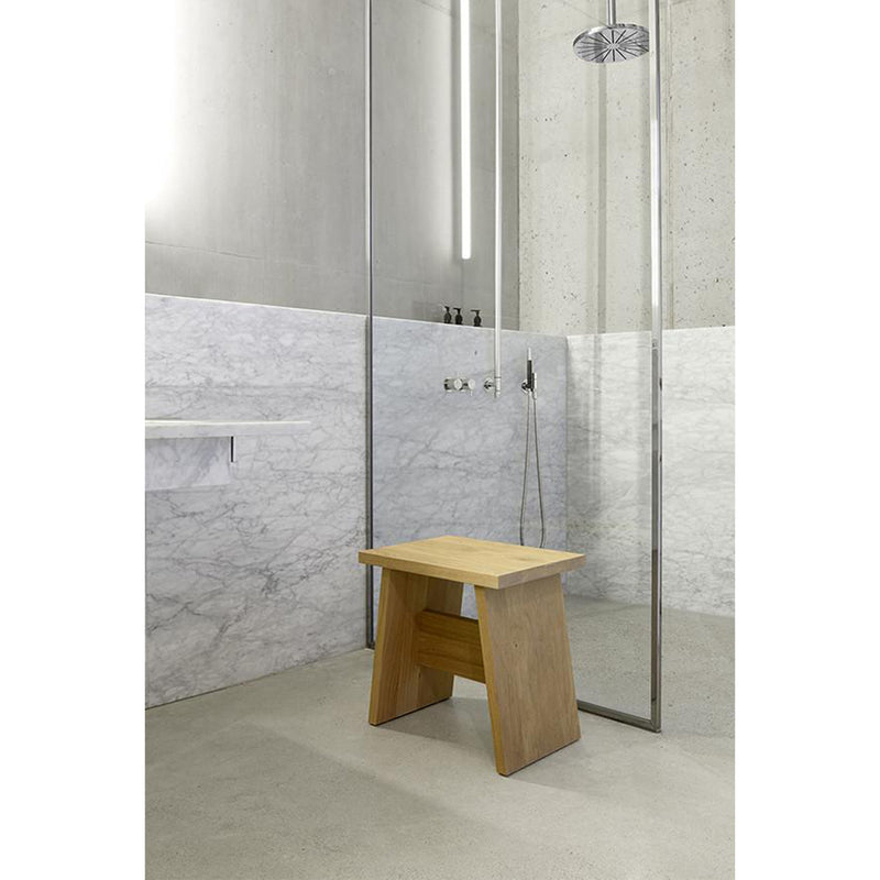 A minimalist bathroom showcases a walk-in shower with a glass partition and ceiling-mounted showerhead. An E15 DC03 LANGLEY stool, crafted from European walnut, rests on the concrete floor, while white marble tiles partially adorn the walls.