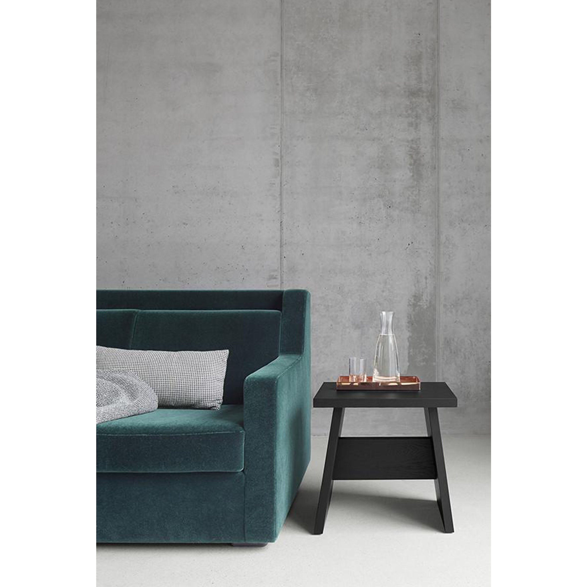 A minimalist living room showcases the E15 DC03 LANGLEY sofa in dark teal beside a modern black side table made of European walnut, which holds a glass carafe and small tray. A light gray concrete wall serves as the backdrop, with a gray pillow resting on the sofa.