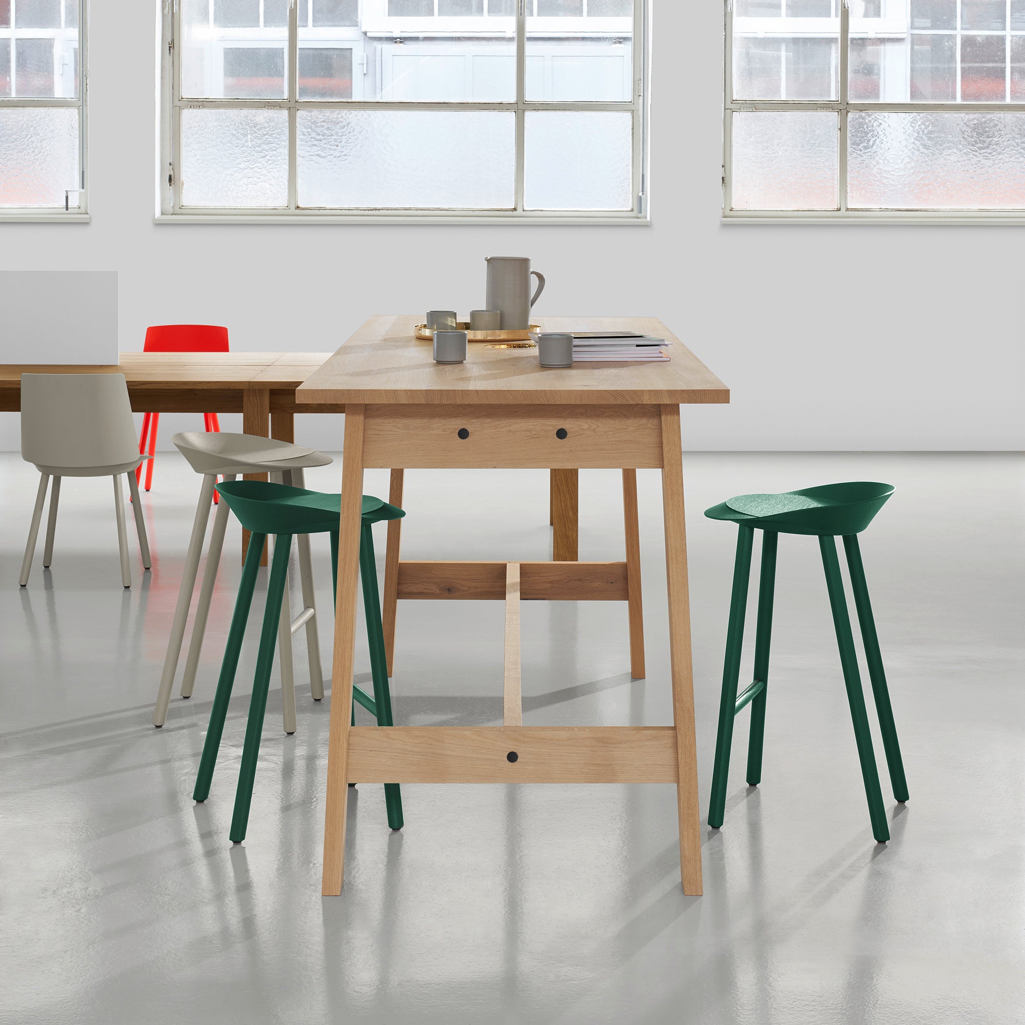 A modern kitchen interior features the DC08 Basis High Table by E15, resembling David Chipperfields style, with green and white stools. The table holds a mug, book, and jar. Surrounding a nearby traditionally crafted dining table are red, white, and green chairs. Large windows flood the space with natural light.