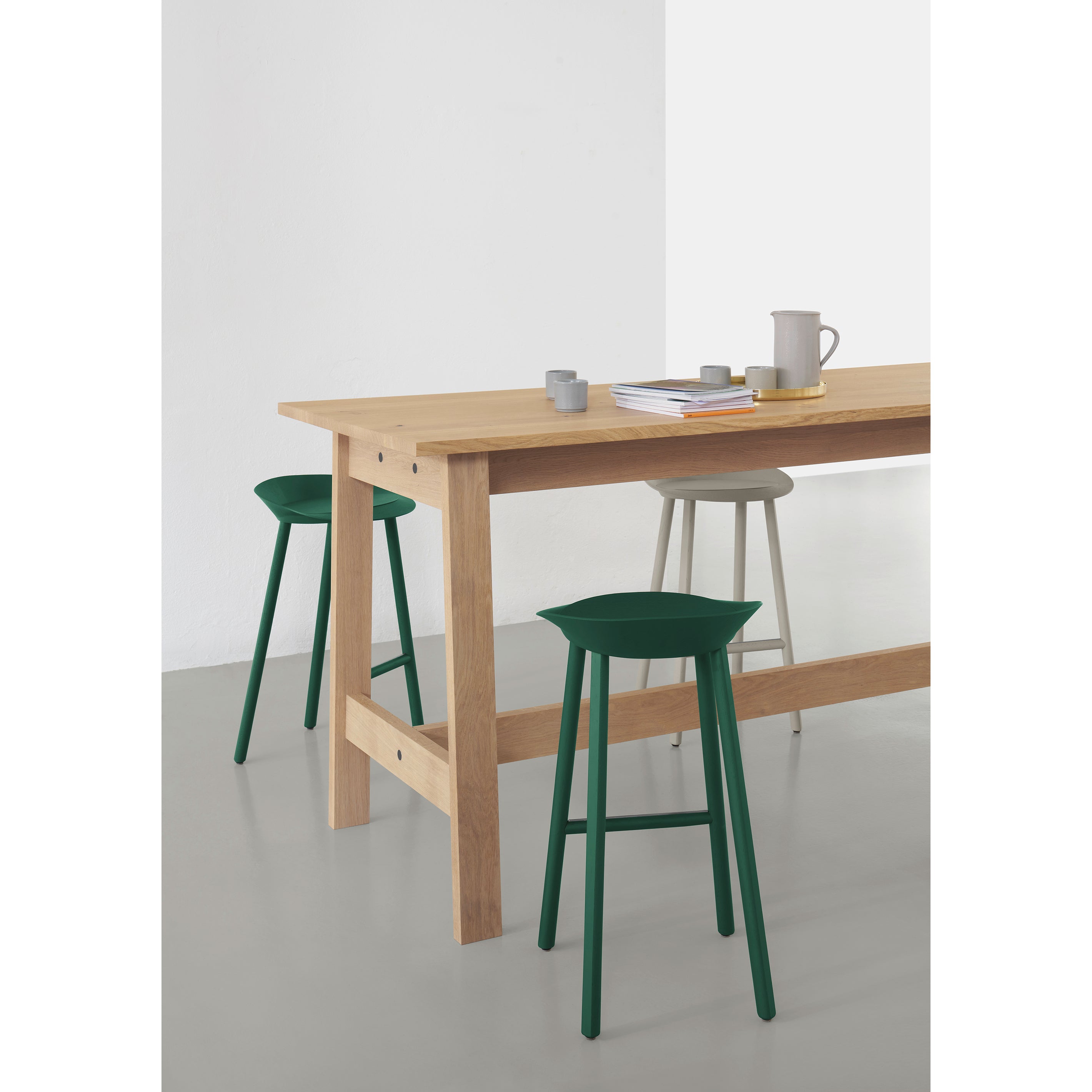 A minimalist dining area highlights the E15 DC08 Basis High Table with green and beige stools. A pitcher, cups, and tray adorn the table. The room’s light grey walls and pale floor reflect David Chipperfields touch, enhancing its serene atmosphere.