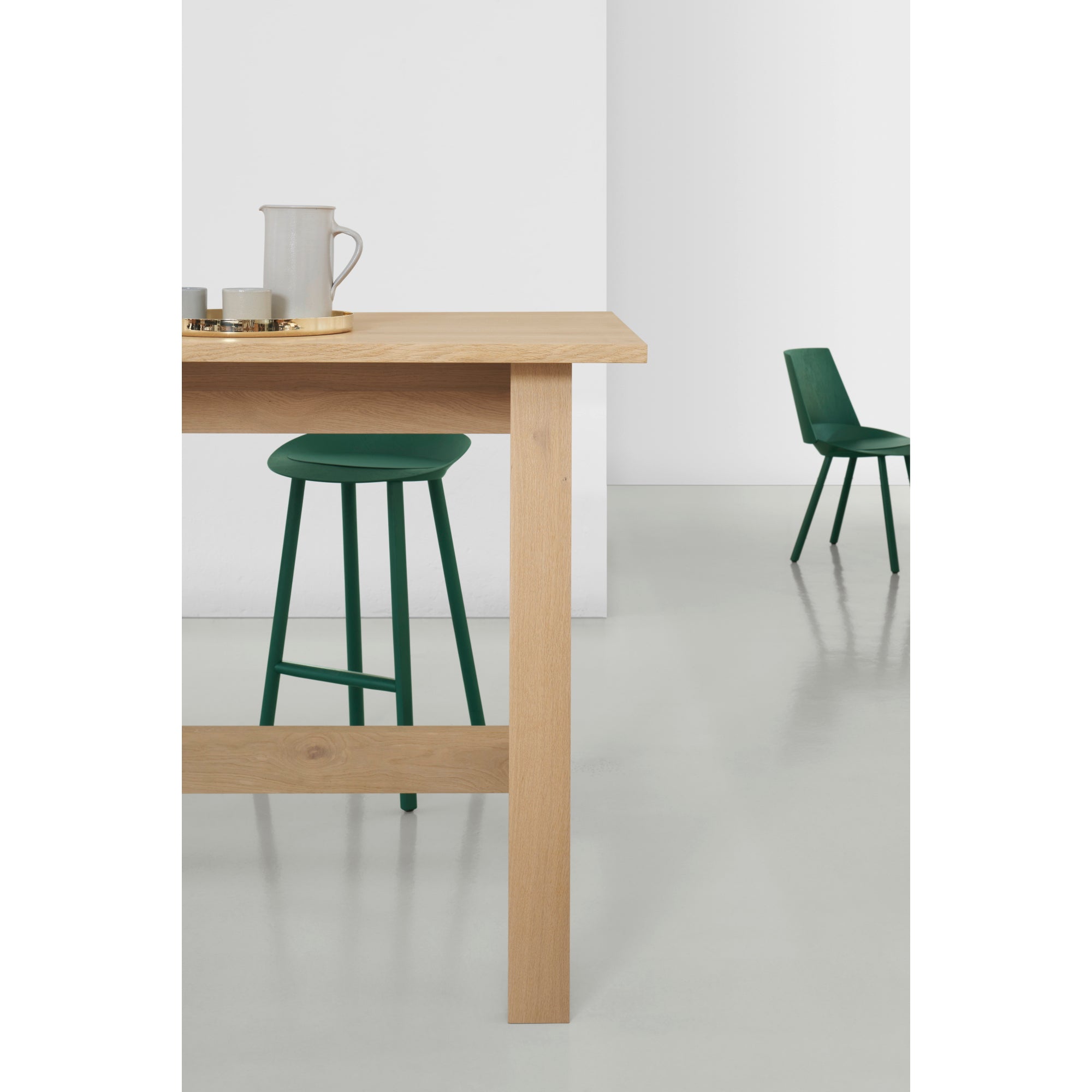 Minimalist room with an E15 DC08 Basis High Table featuring a tray with a pitcher and cups. A green barstool fits beneath. In the background, matching green seating reflects David Chipperfields style against a light floor and white wall.