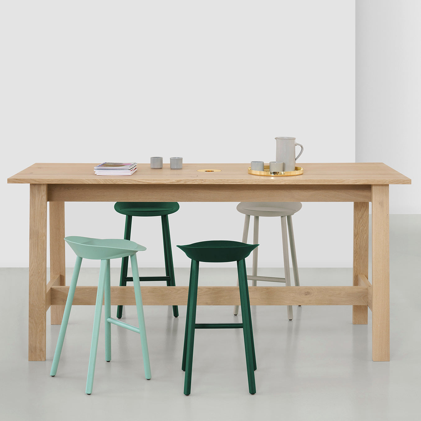 The E15 DC08 Basis High Table, with minimalist design and sturdy construction, is complemented by four stools in pastel green, dark green, and beige. The tables light surface hosts small candles, a tray, a pitcher, and stacked bowls.