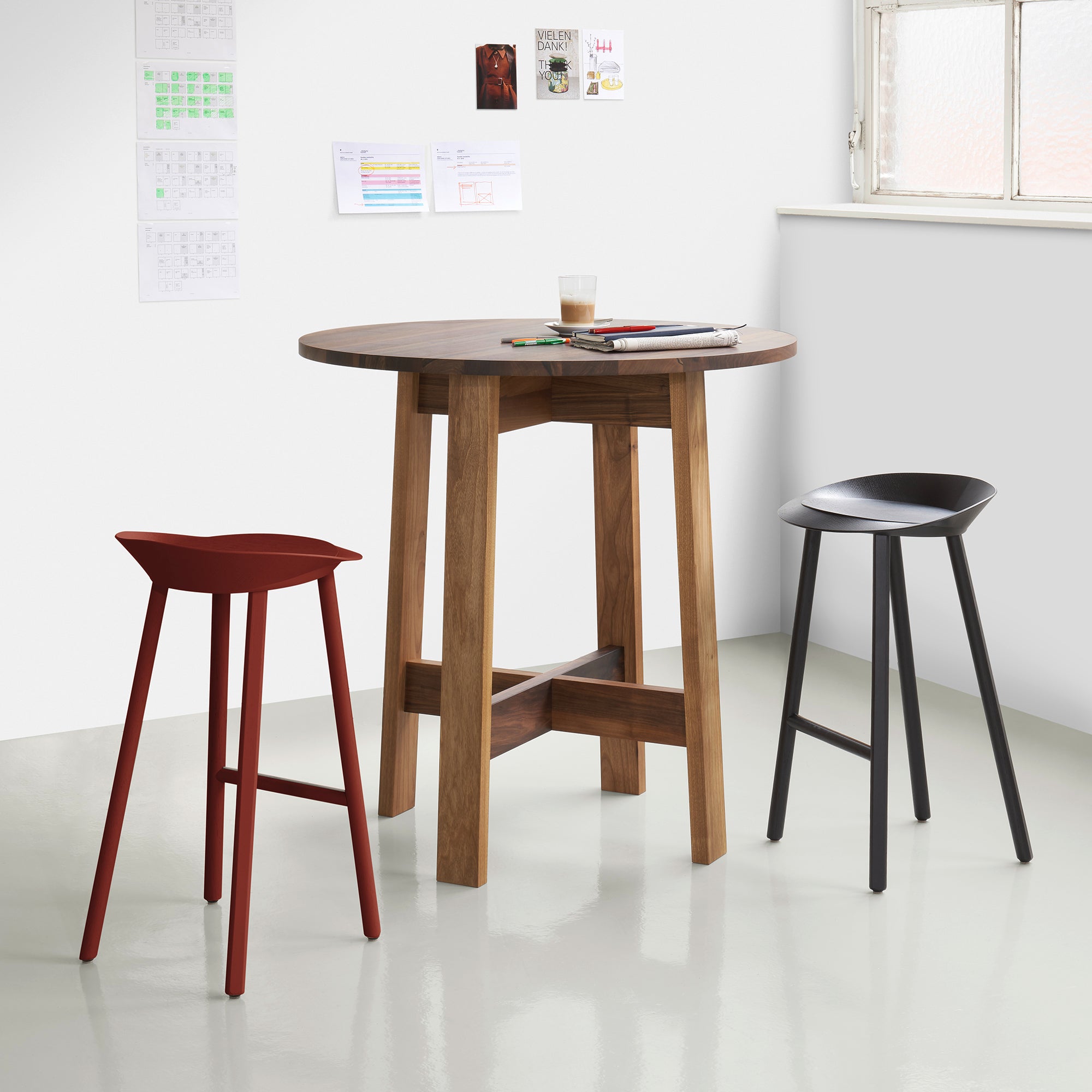 A modern room boasts an E15 DC08 Basis High Table Round with two bar stools: one red with a curved seat and the other black with a backrest. White walls adorned with papers and artwork, along with a window on the right, allow natural light in.
