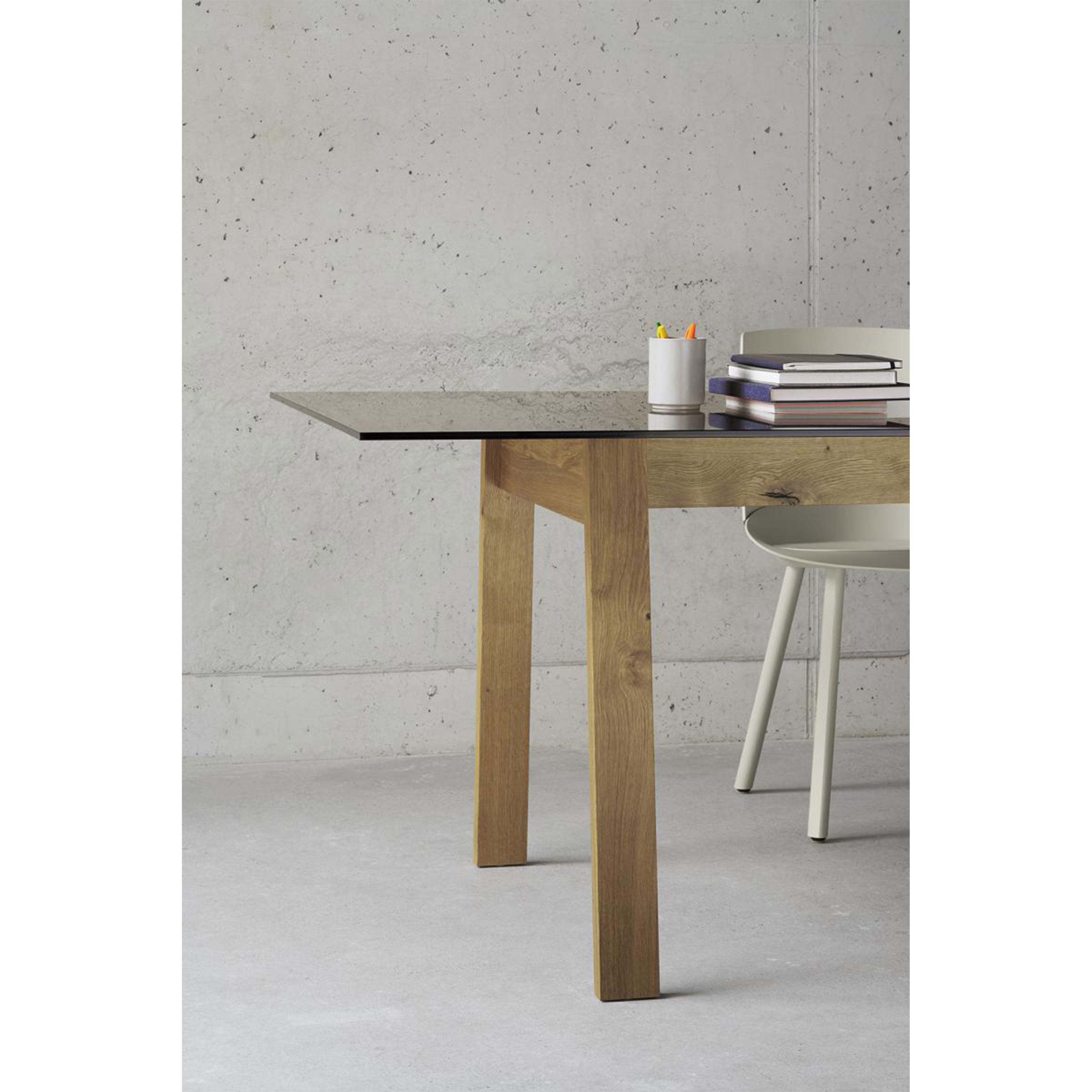 A minimalist study space features an E15 DC08 Basis Table with a glass top, supported by solid wood trestles. A beige chair sits against a concrete wall backdrop, while the table displays a white cup of colored pencils and a stack of books.