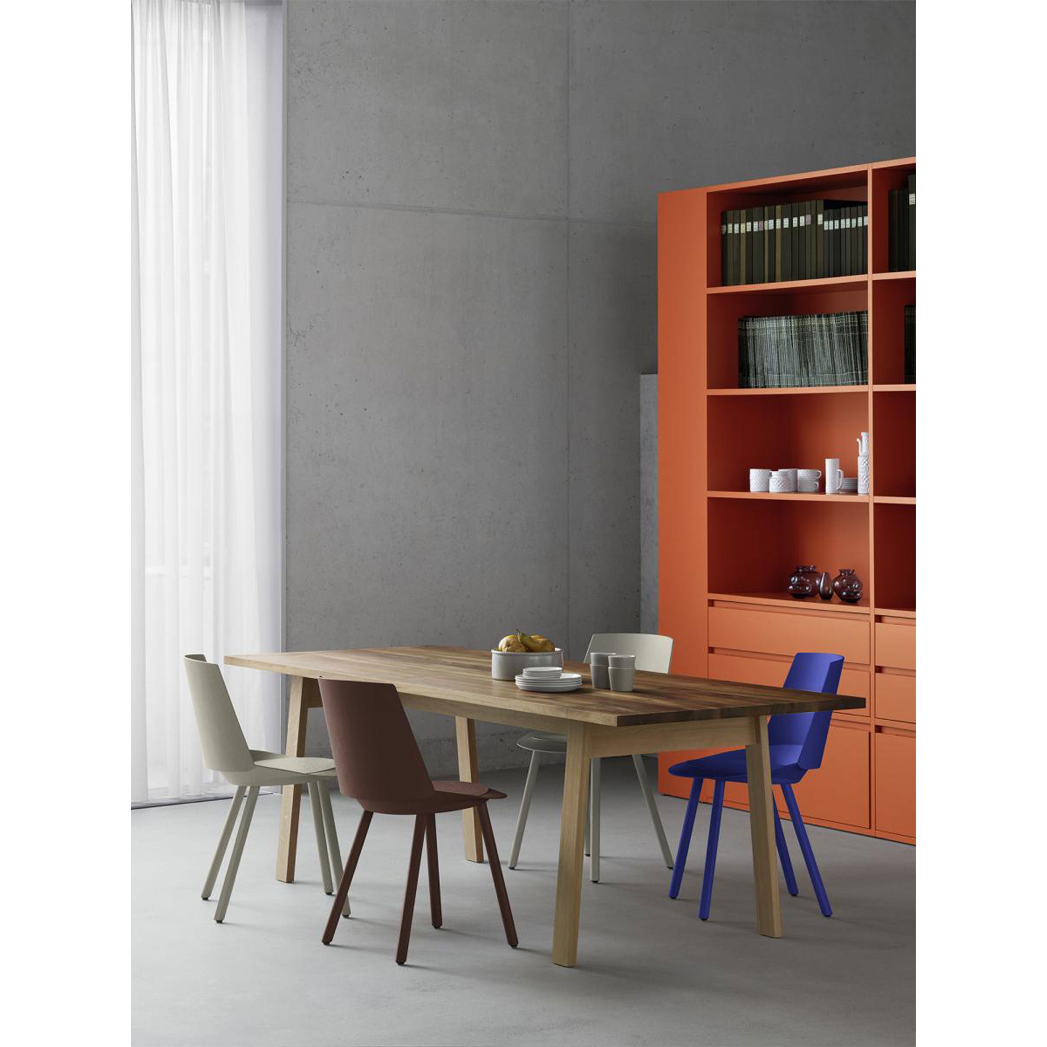 The minimalist dining room showcases an E15 DC08 Basis Table with its solid wood trestles, surrounded by mismatched chairs in neutral and vibrant colors. An orange shelving unit brightens the space while sheer white curtains add a soft touch.