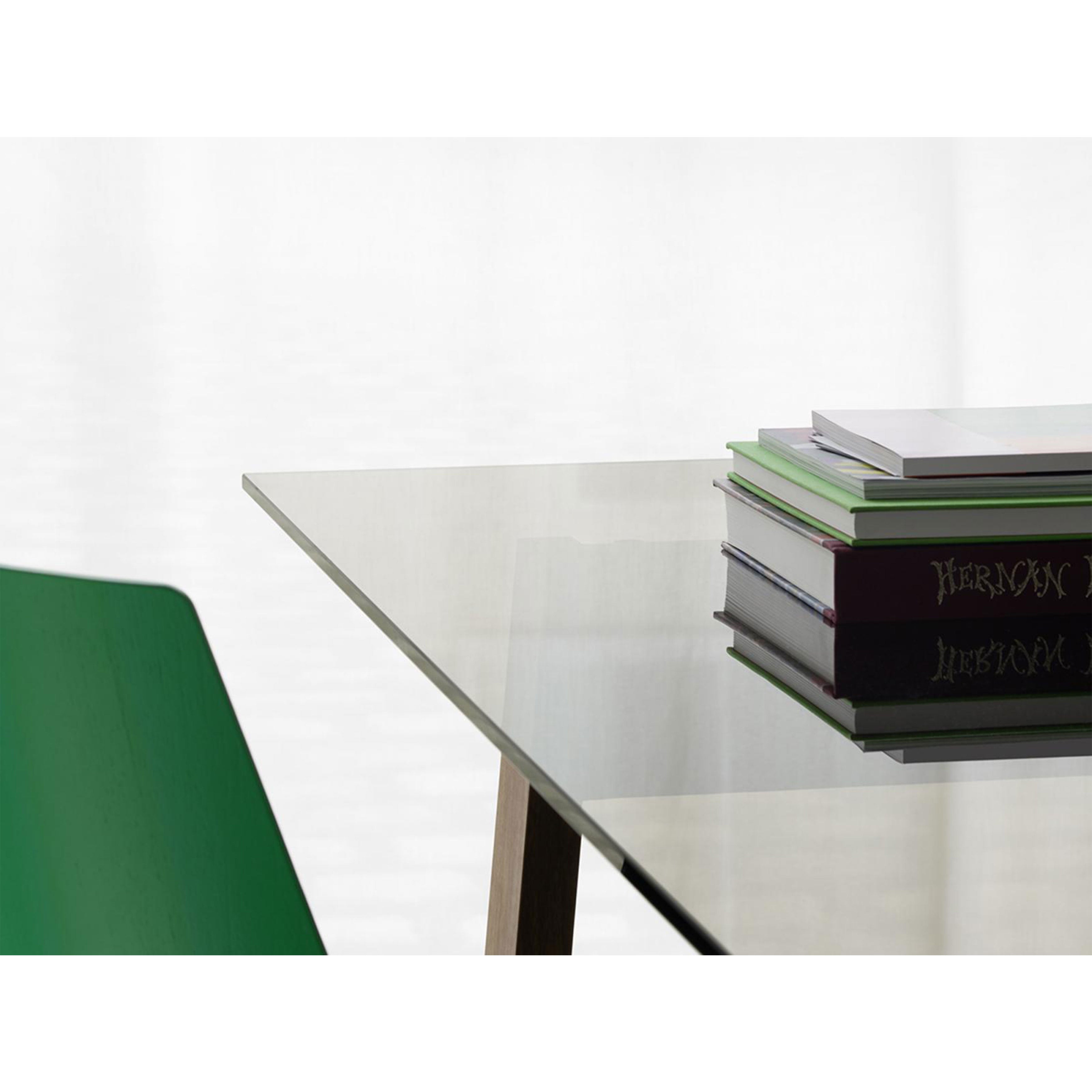 An E15 DC08 Basis Table with a glass top holds a stack of books in one corner. Soft lighting illuminates the scene, where part of a green chair and solid wood trestles provide warmth and charm.