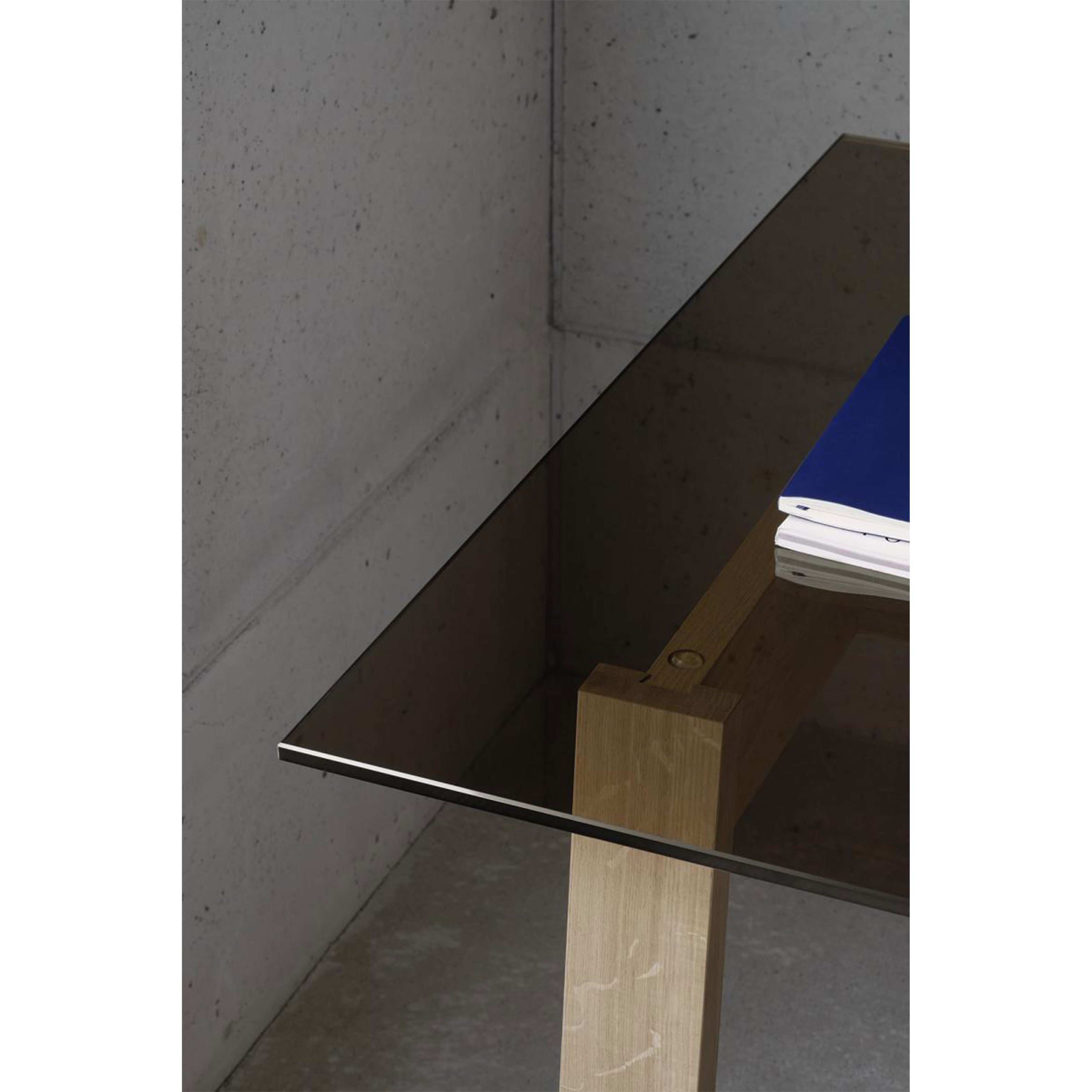 A close-up of the sleek DC08 Basis Table by E15, featuring a transparent glass top and solid wood trestles. A stack of blue and white papers rests on the tables edge against a plain, gray concrete wall.