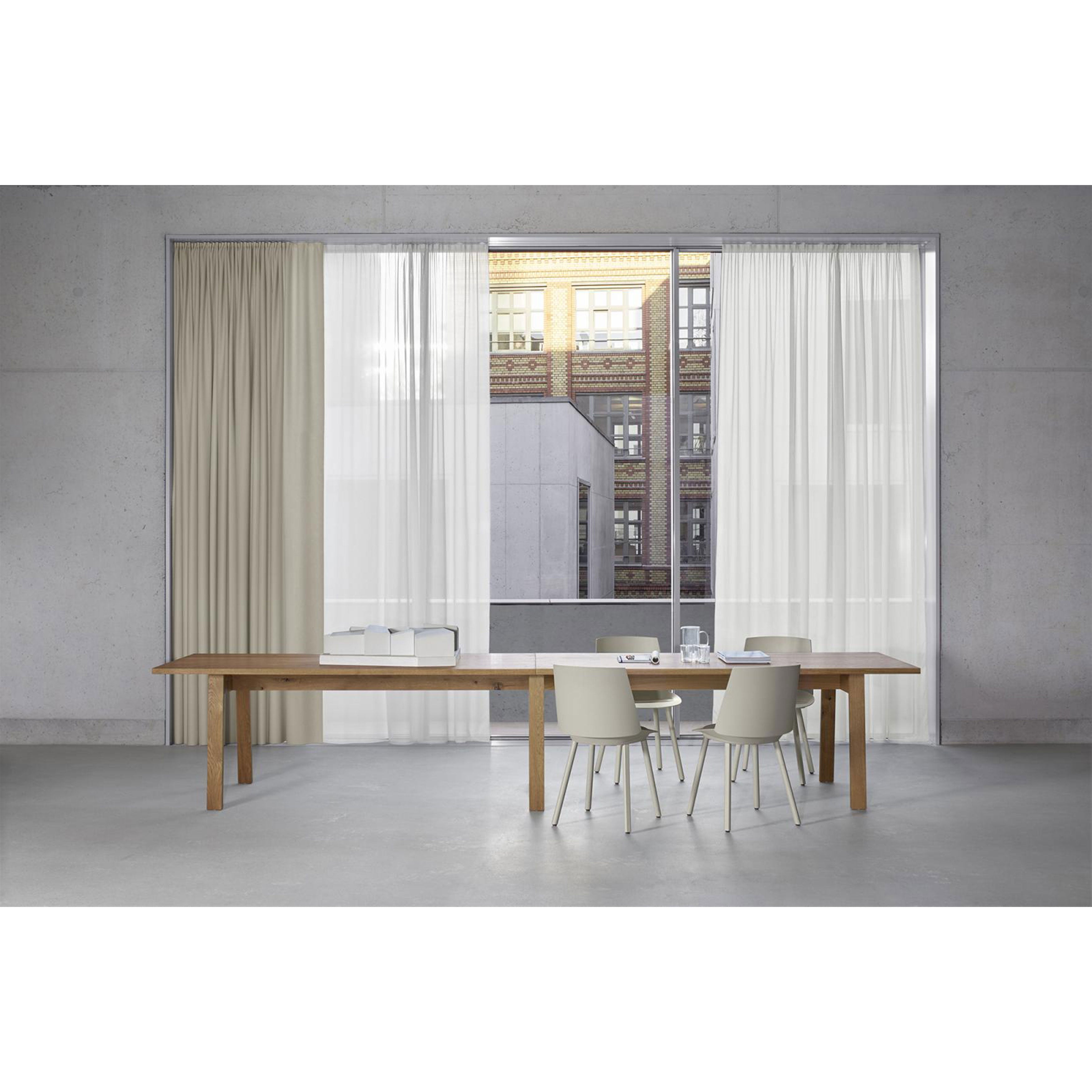 A minimalist dining area showcases the E15 DC08 Basis Table with solid wood trestles, surrounded by four white chairs. Large windows with beige and white curtains reveal a view of a brick and concrete building. Gray floors and walls enhance the rooms clean, modern look.