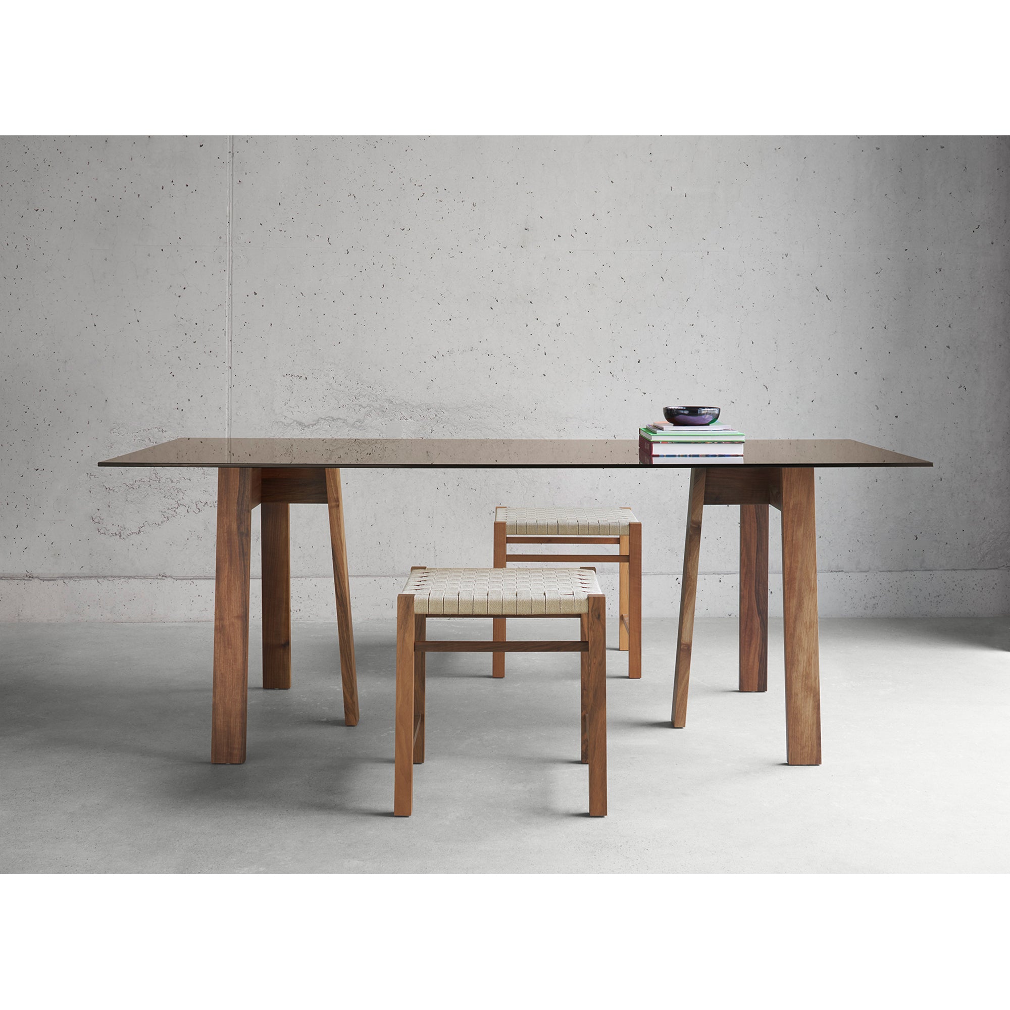 A minimalist dining setup includes the DC08 Basis Trestle by E15, featuring solid wood trestles and two matching chairs with woven seats. A small stack of books and a bowl rest on the table, against a plain light gray wall.