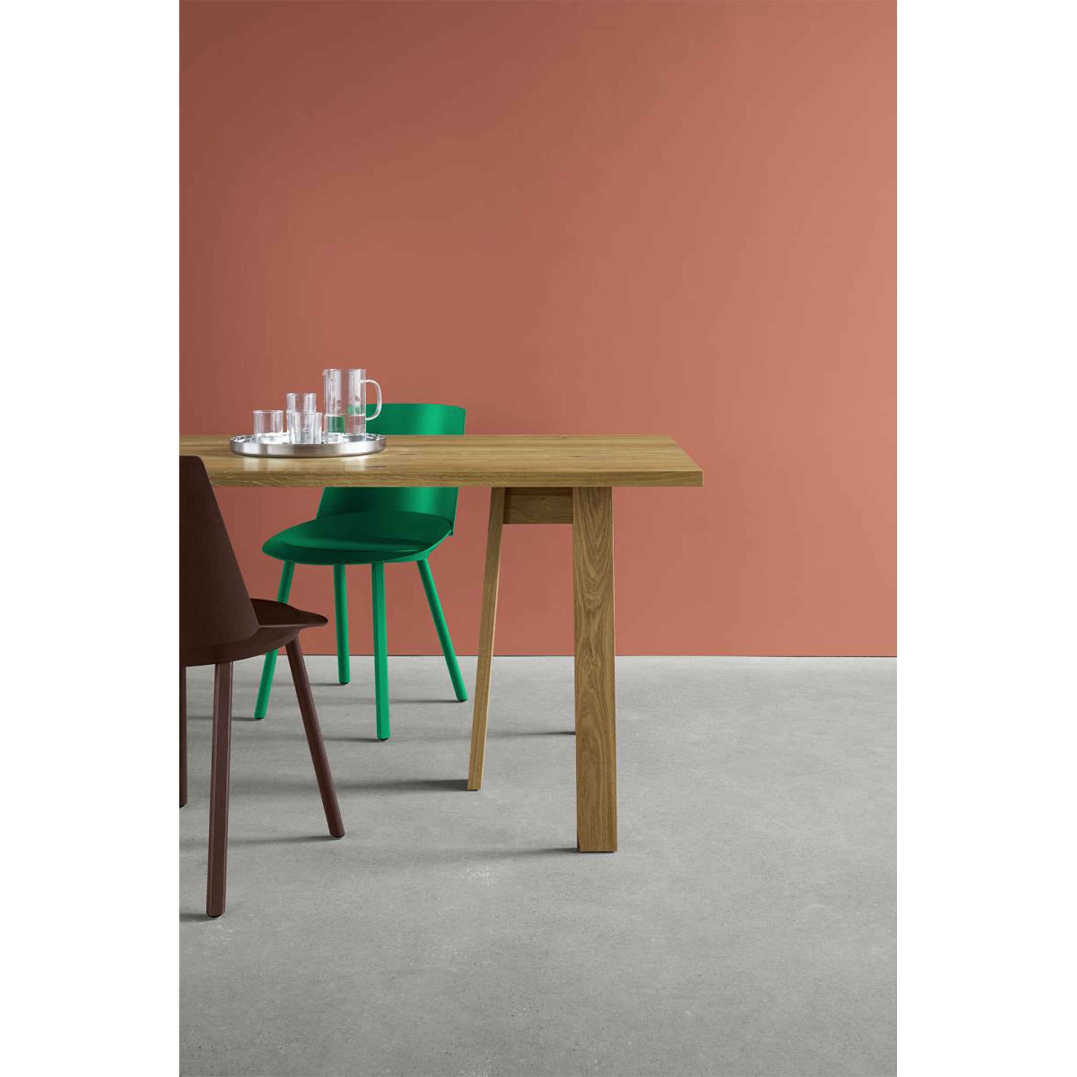 In a minimalist room, an E15 DC08 Basis Trestle table with solid wood trestles is adorned with a tray holding a pitcher and cups. A green chair and a brown chair are arranged around it, set against terracotta-colored walls and a light gray floor.