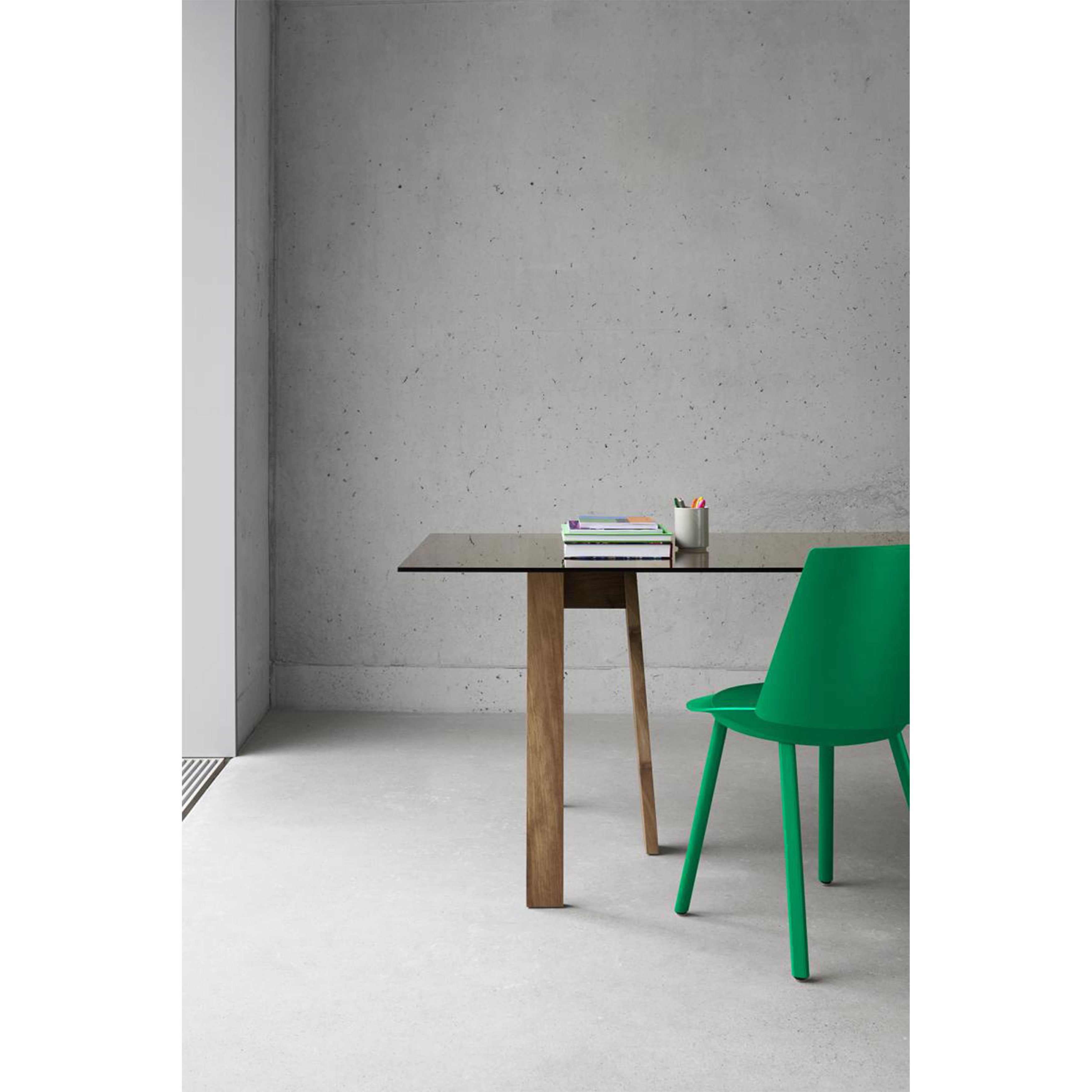 A minimalist interior showcases a concrete wall and floor. An E15 DC08 Basis Trestle with a glass top holds books and colored pencils. A green chair sits beside the table, combining simplicity with modern flair.