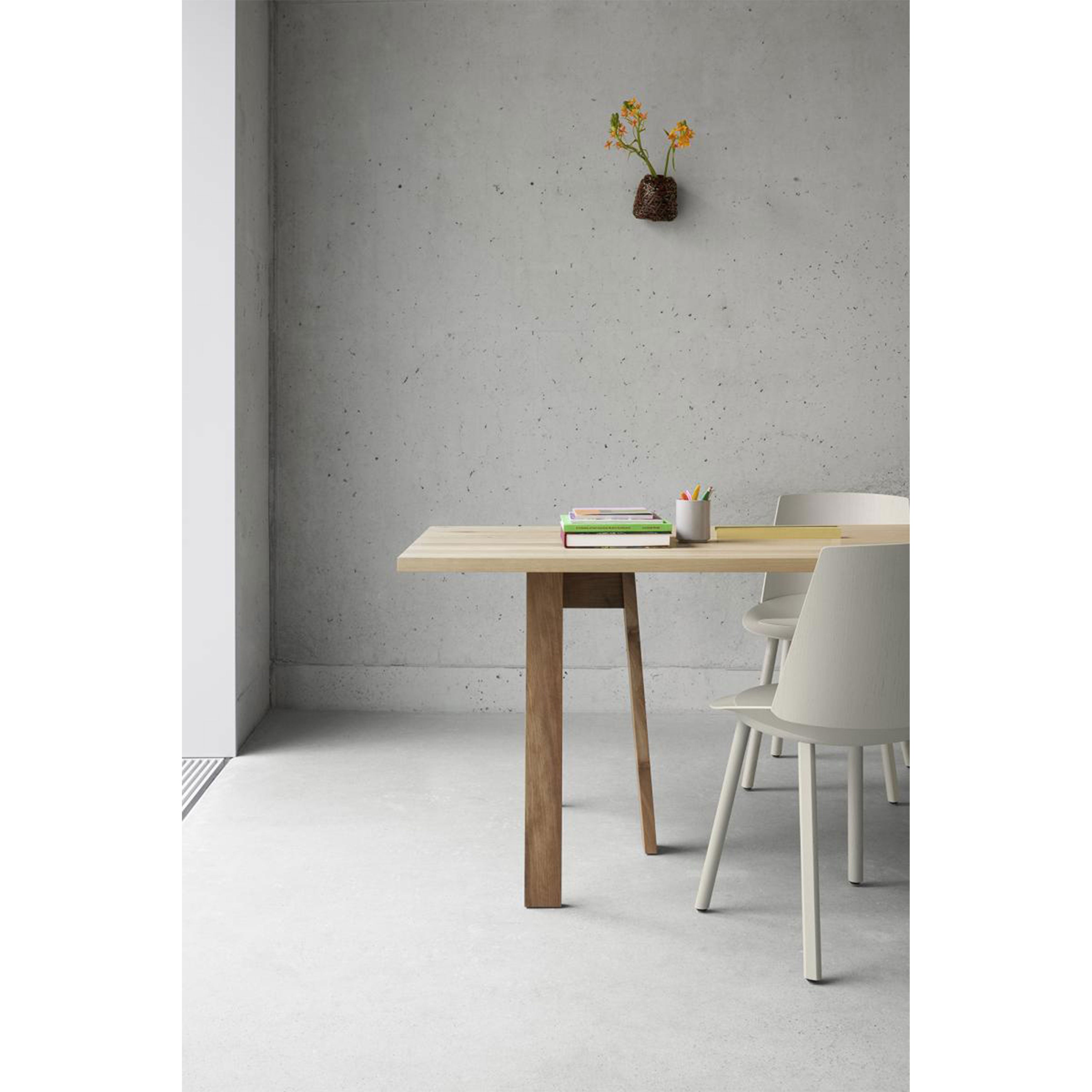 The minimalist room features an E15 DC08 Basis Trestle table in light wood, accompanied by two white chairs. A concrete wall and floor provide the backdrop, enhanced by a vase of yellow flowers. Bathed in natural left light, the table also holds a stack of books and a white pencil holder.
