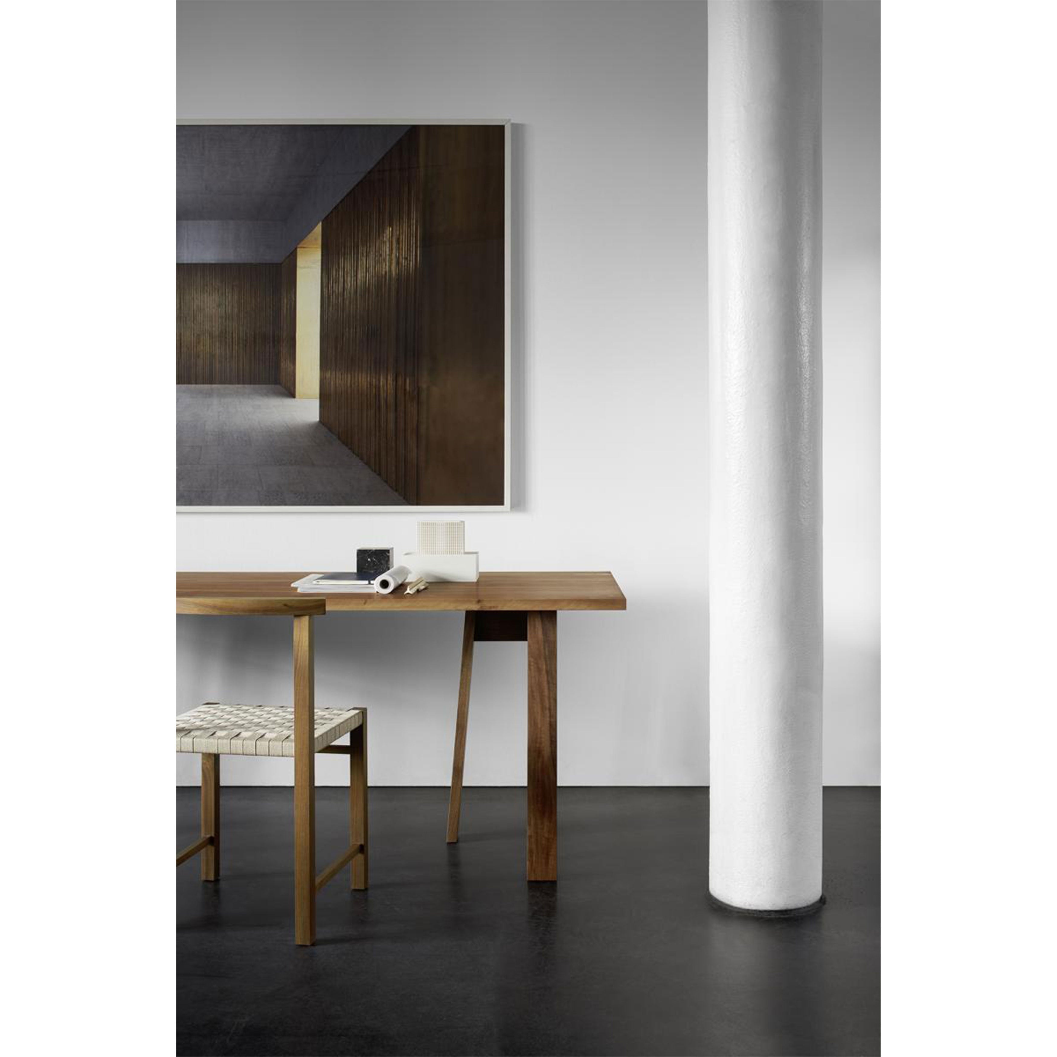 A minimalist interior features an E15 DC08 Basis Trestle table on solid wood legs, complemented by a woven chair on a dark floor. A large abstract painting decorates the wall above, while a tall white column on the right enhances the clean, modern aesthetic.