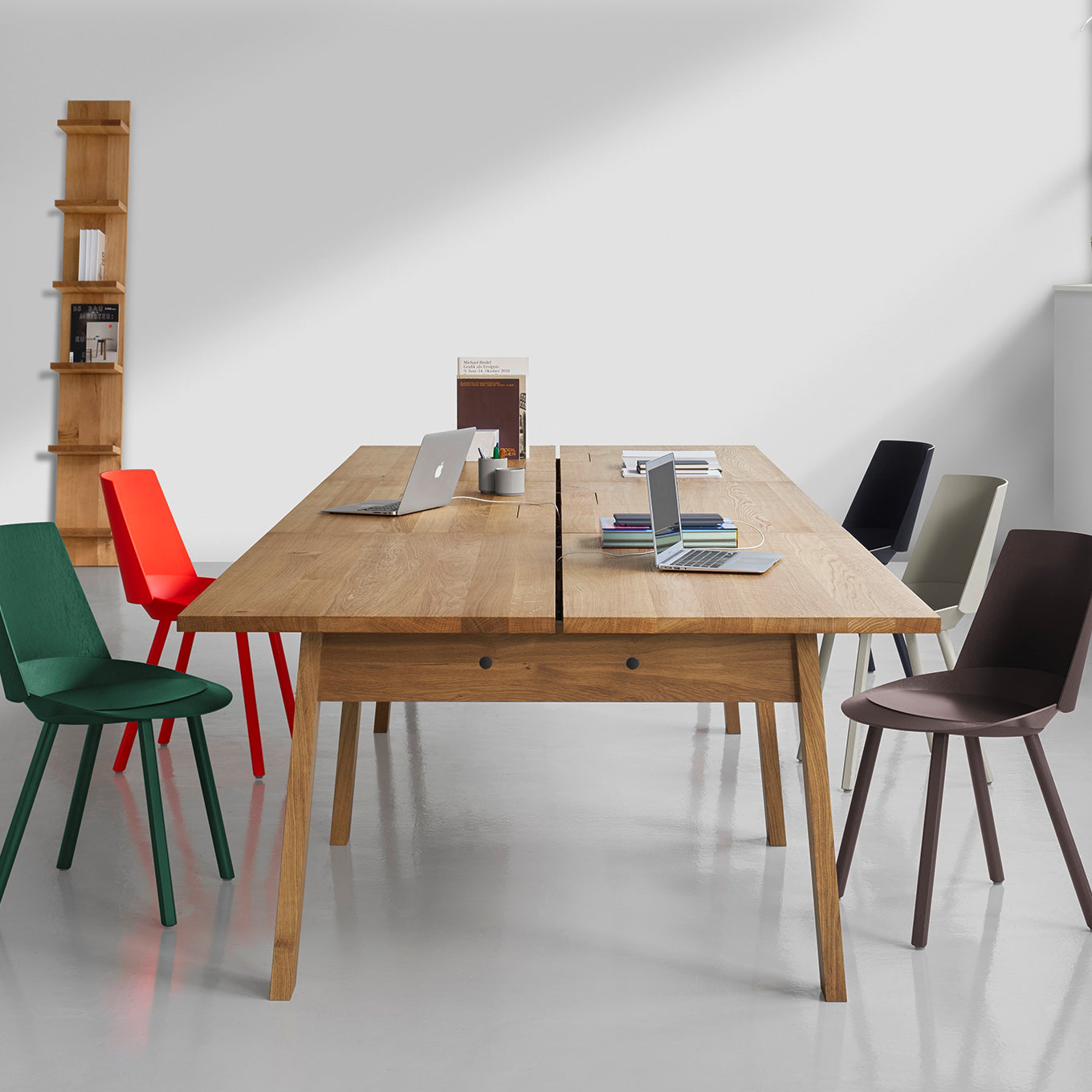A modern office features an E15 DC08 Basis Workstation with a long wooden table, vibrant chairs, and laptops on top. A small book-filled shelf against the white wall perfectly complements this creative work environment.