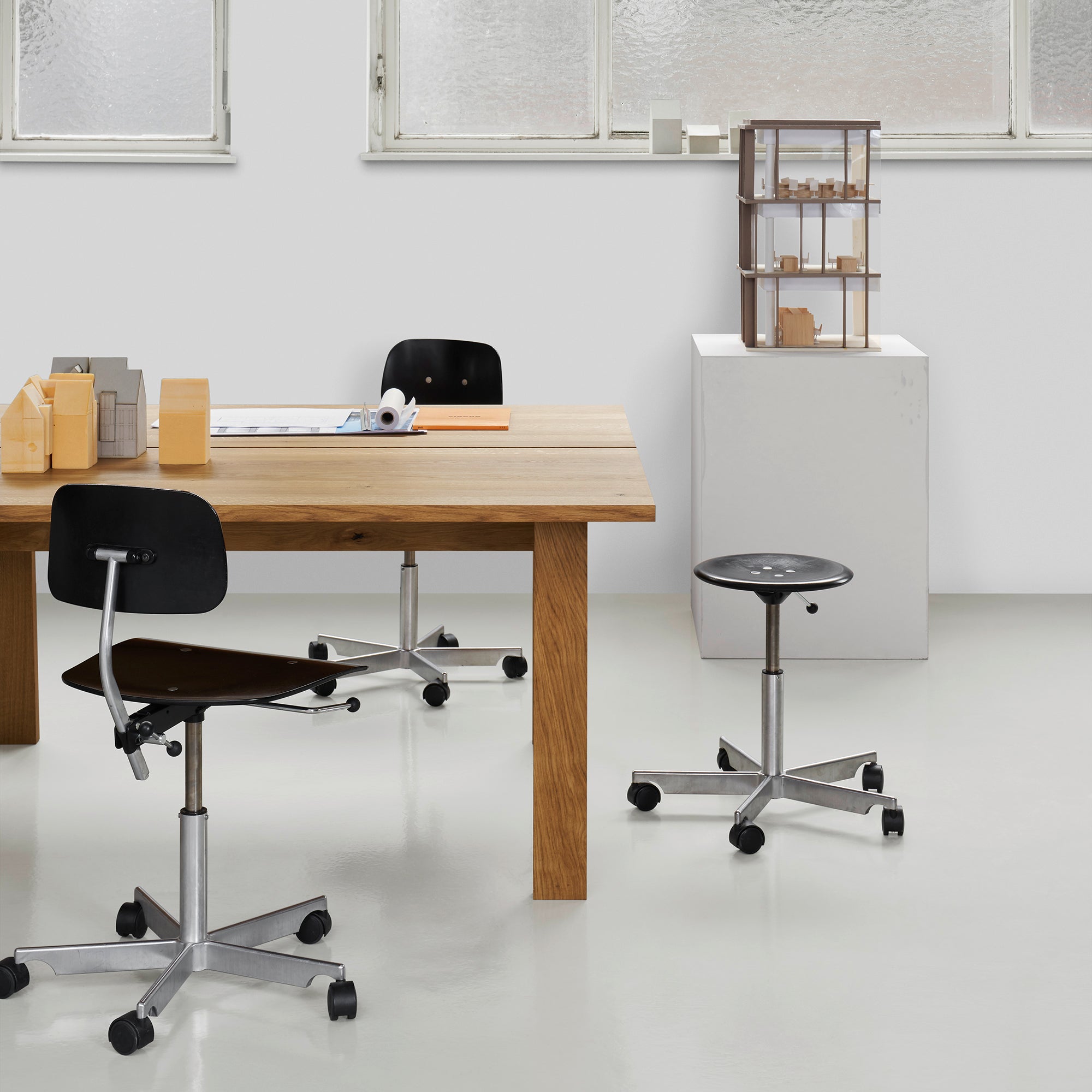 A modern office showcases an E15 DC08 Basis Workstation with a wooden desk, two adjustable black chairs with metal bases, and a small wheeled stool. A white pedestal display case is set against large frosted windows in a white wall, creating an inspiring and creative workspace.