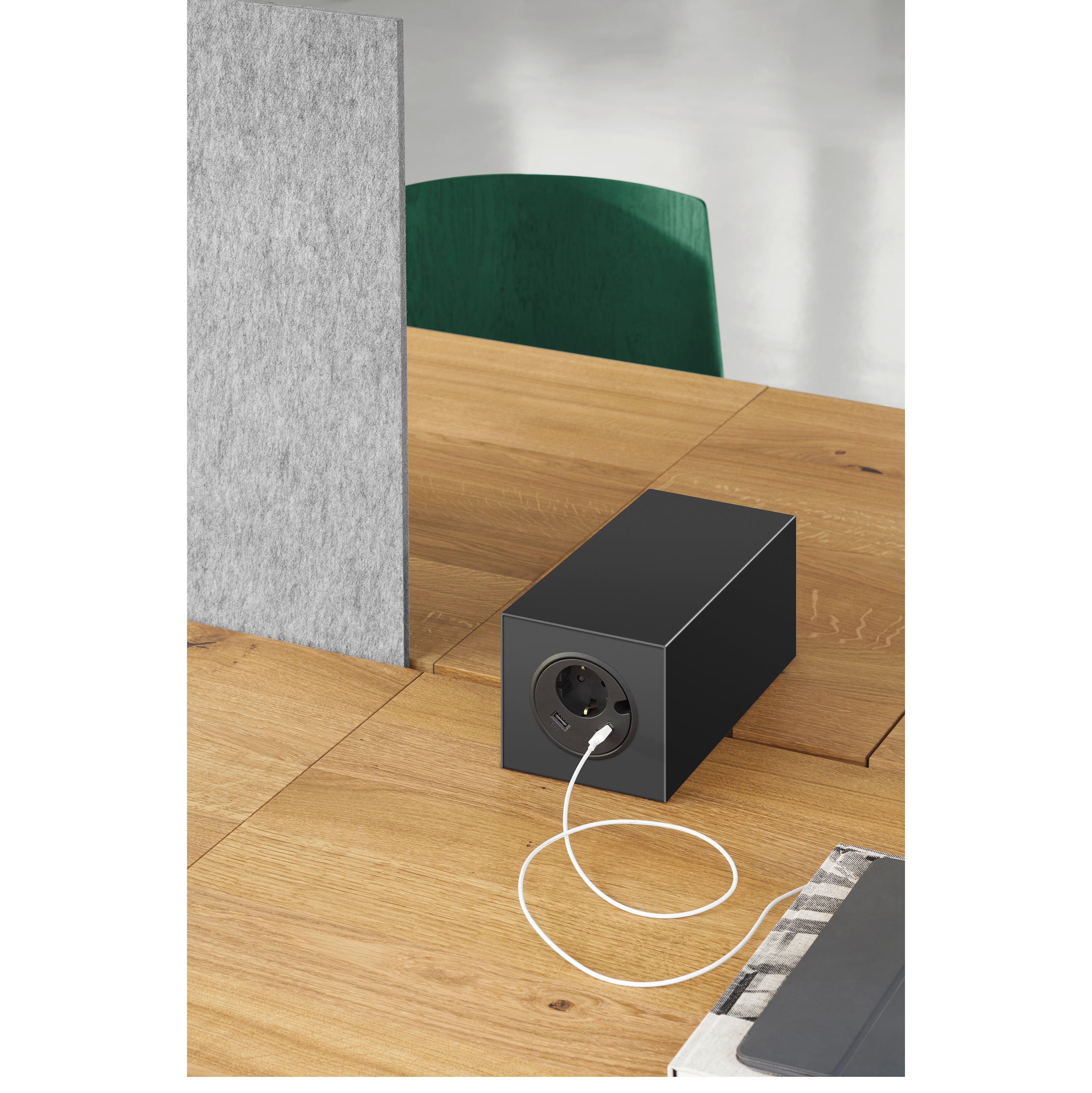 The E15 DC08 Basis Workstation is a modern wooden table with a black power hub, connected by a white cable for enhanced functionality. A gray partition and green chair in the background complete this creative work environment.