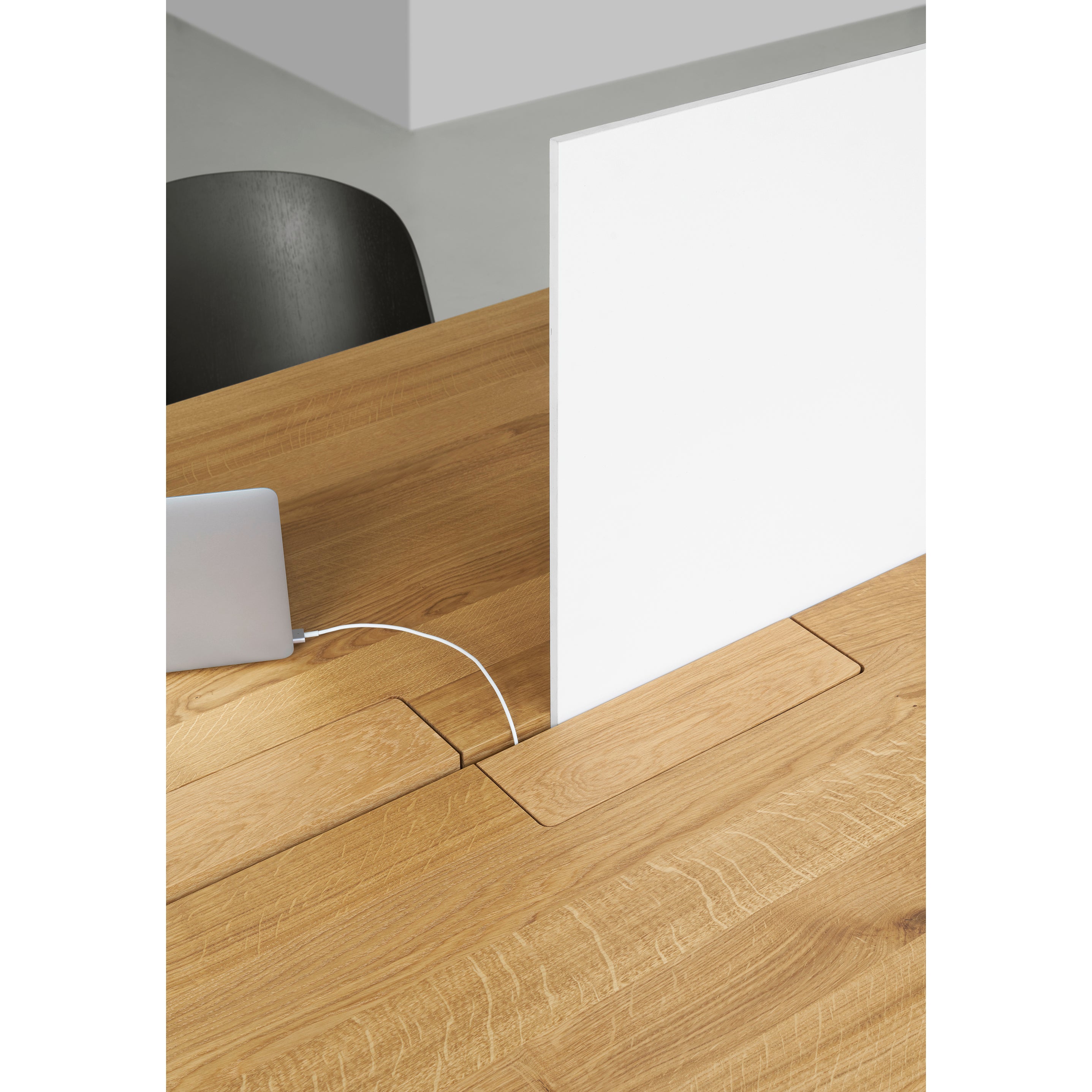 A modern E15 DC08 Basis Workstation includes a wooden table with a cable management slot, a partially visible laptop charger, and a white partition screen. A black chair is slightly visible in the creative work environment.