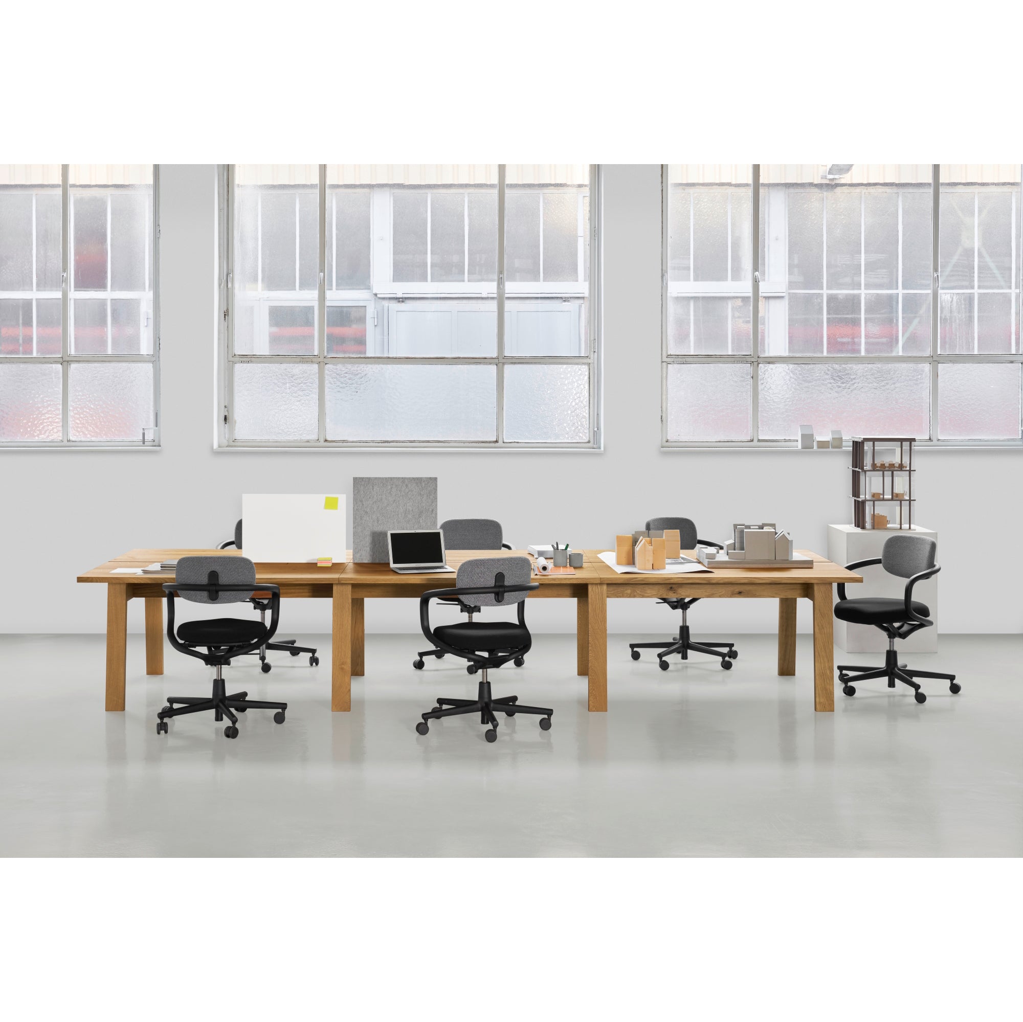 The spacious office features an E15 DC08 Basis Workstation with three wooden desks, each paired with office chairs, computers, and various supplies. Large windows provide natural light, creating a minimalistic and organized creative work environment.