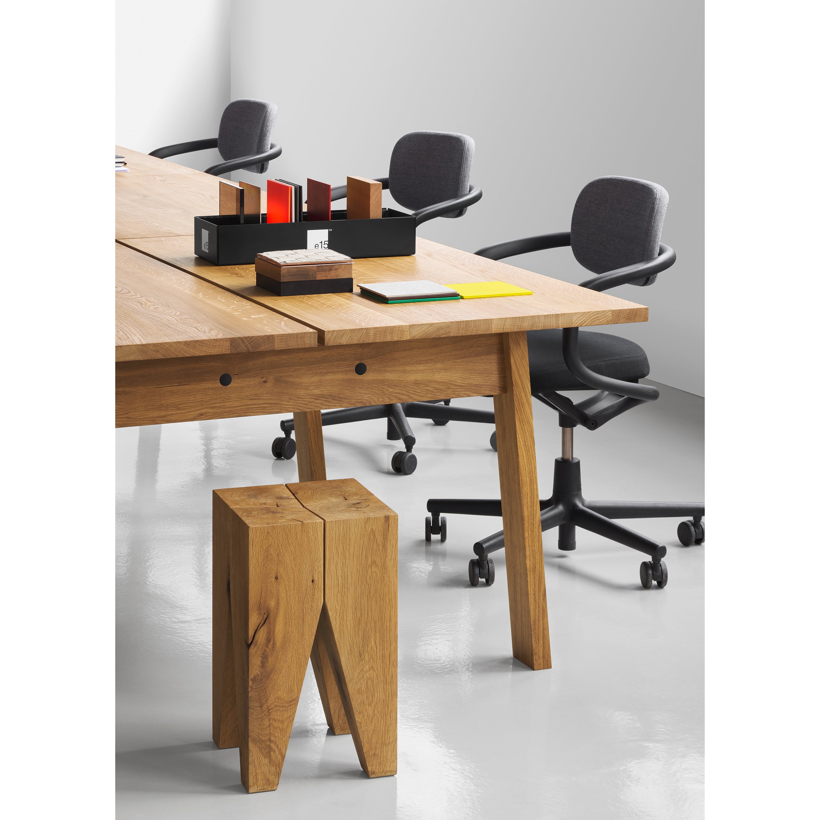 A modern office has an E15 DC08 Basis Workstation featuring a wooden table with a matching wooden stool, three black swivel chairs on a shiny white floor, and a black organizer with colorful files. The setup is against a plain white wall.