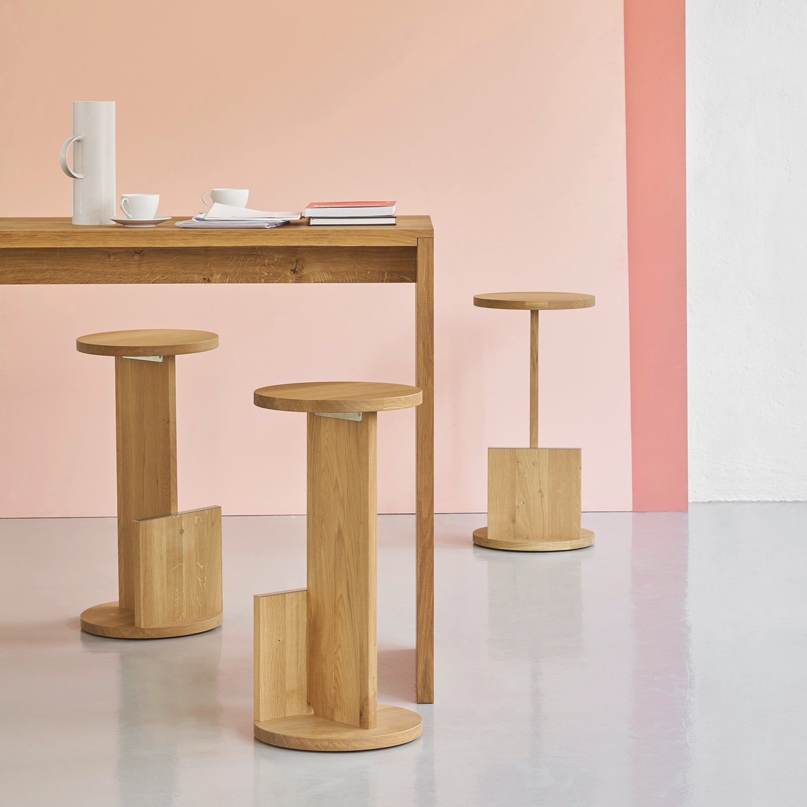 A modern dining area features a solid European oak table with three E15 DT01 Tank Barstools. A gradient wall transitions from pale pink to white, while a minimalist setup includes a white pitcher and teacups, reflecting David Thulstrups design ethos.