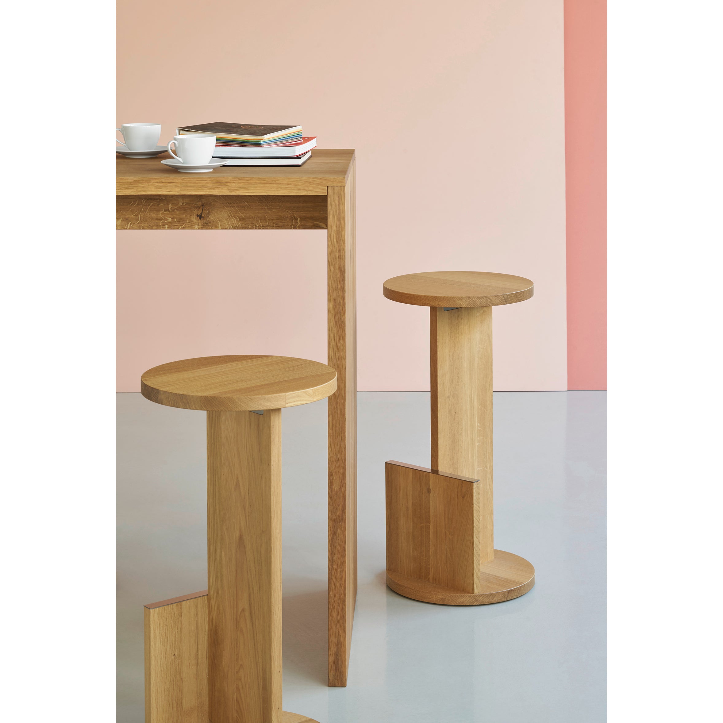 Minimalist DT01 Tank Barstool and table by E15, crafted from solid European oak, sit against a pastel pink wall. The table holds two white cups and books. The bright, modern setting with a smooth white floor reflects David Thulstrups design philosophy.
