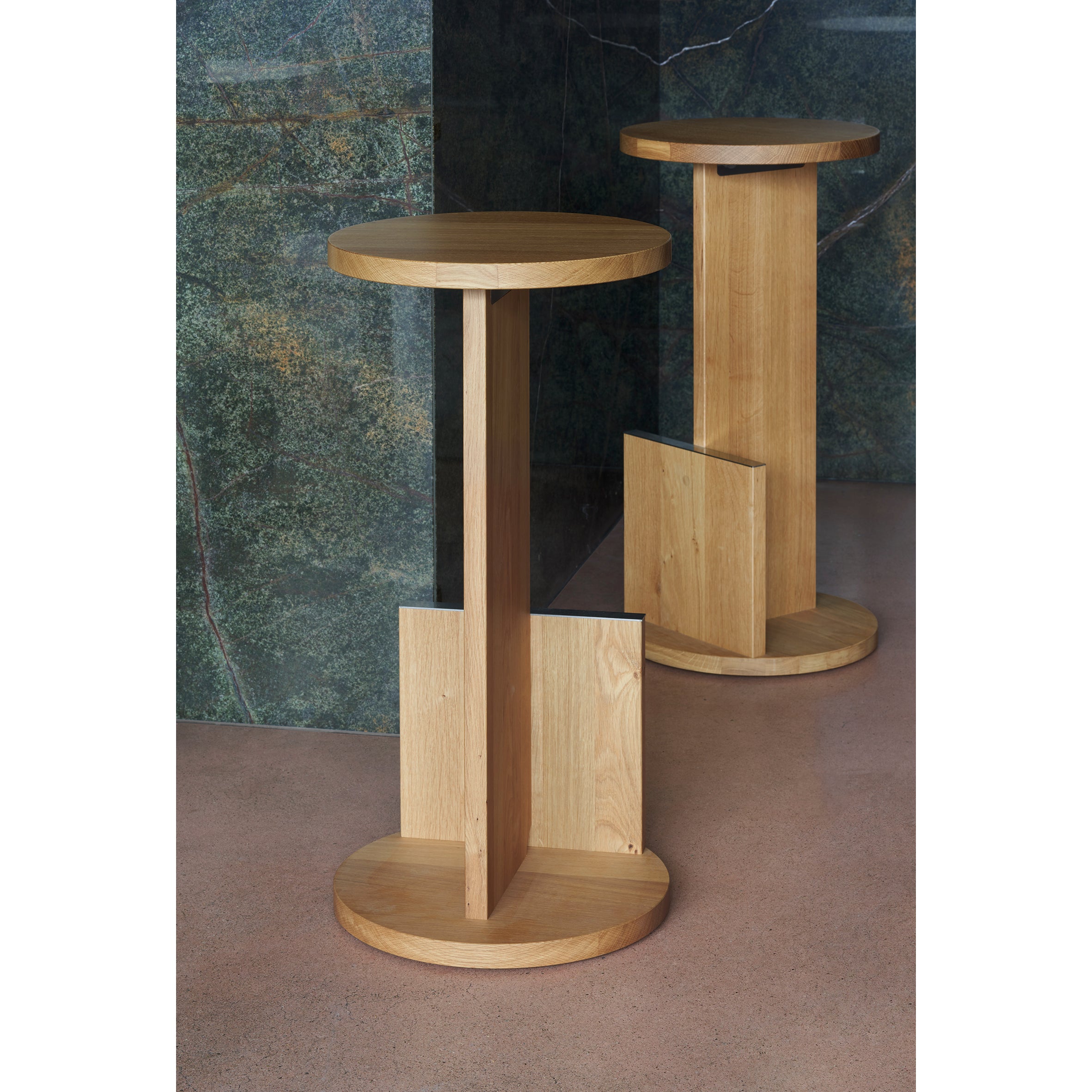 The DT01 Tank Barstool by E15, crafted from solid European oak with round tops and geometric bases, rests on a polished concrete floor. Reminiscent of David Thulstrups designs, they enhance the minimalist elegance against a green marbled wall.