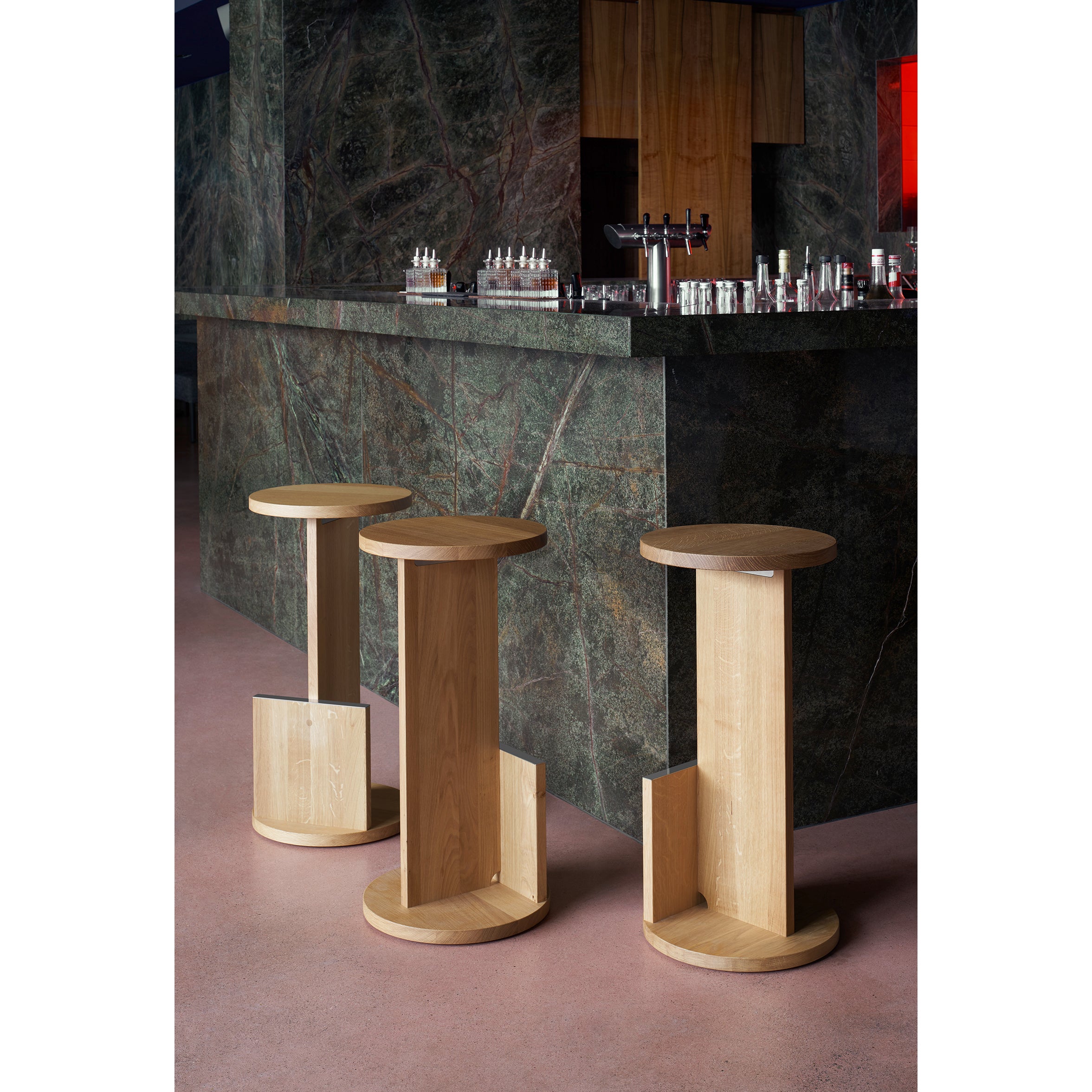 Three DT01 Tank Barstools by E15, crafted from solid European oak, adorn a marble bar counter designed by David Thulstrup. The counter, with subtle greenish hues and sleek bar equipment, is complemented beautifully by the smooth pink-toned floors minimalist elegance.