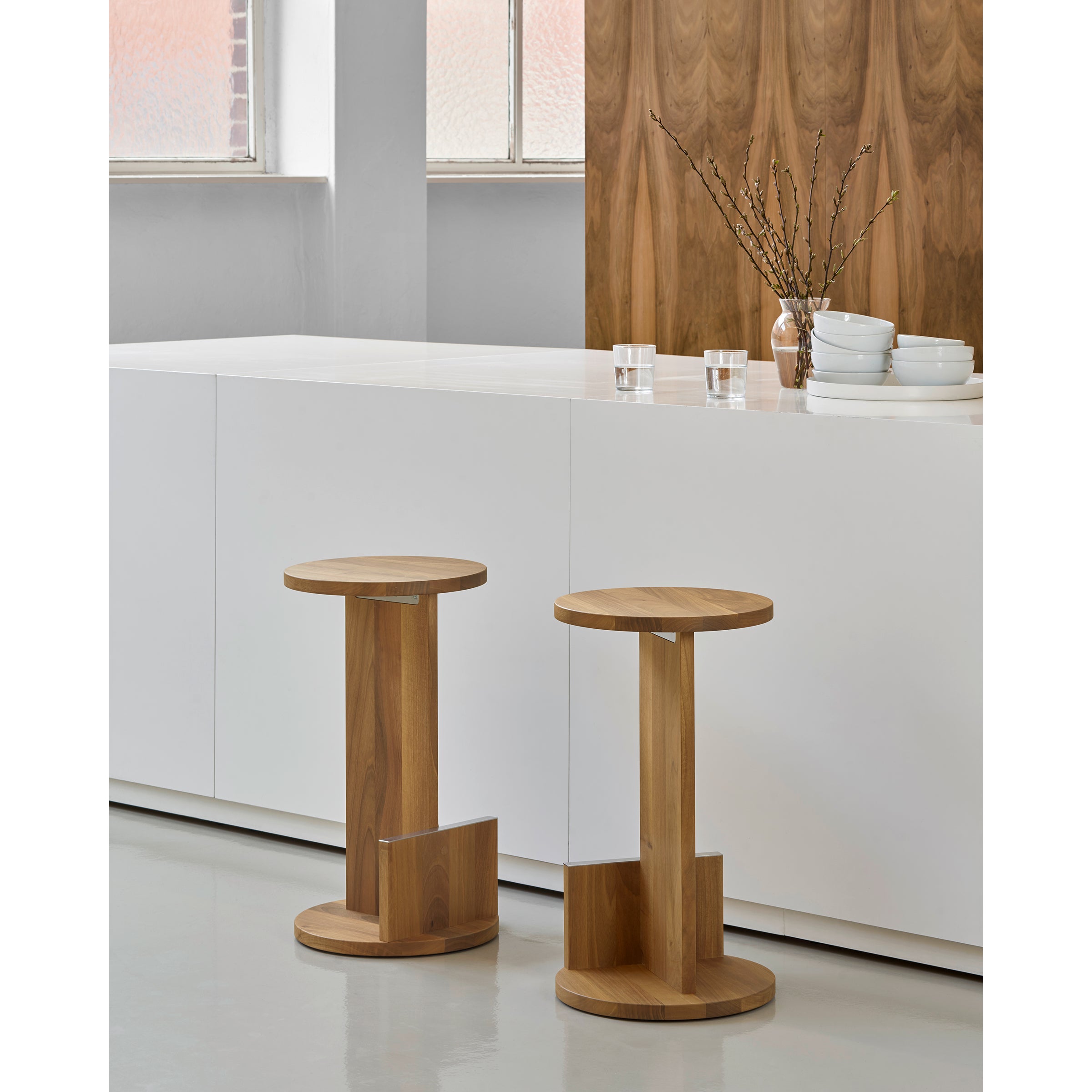 Two DT01 Tank Barstools by E15, crafted from solid European oak, feature circular seats and bases near a white counter with stacked white bowls, cups, and a clear vase holding branches. A large window and wood-paneled wall designed by David Thulstrup are in the background.