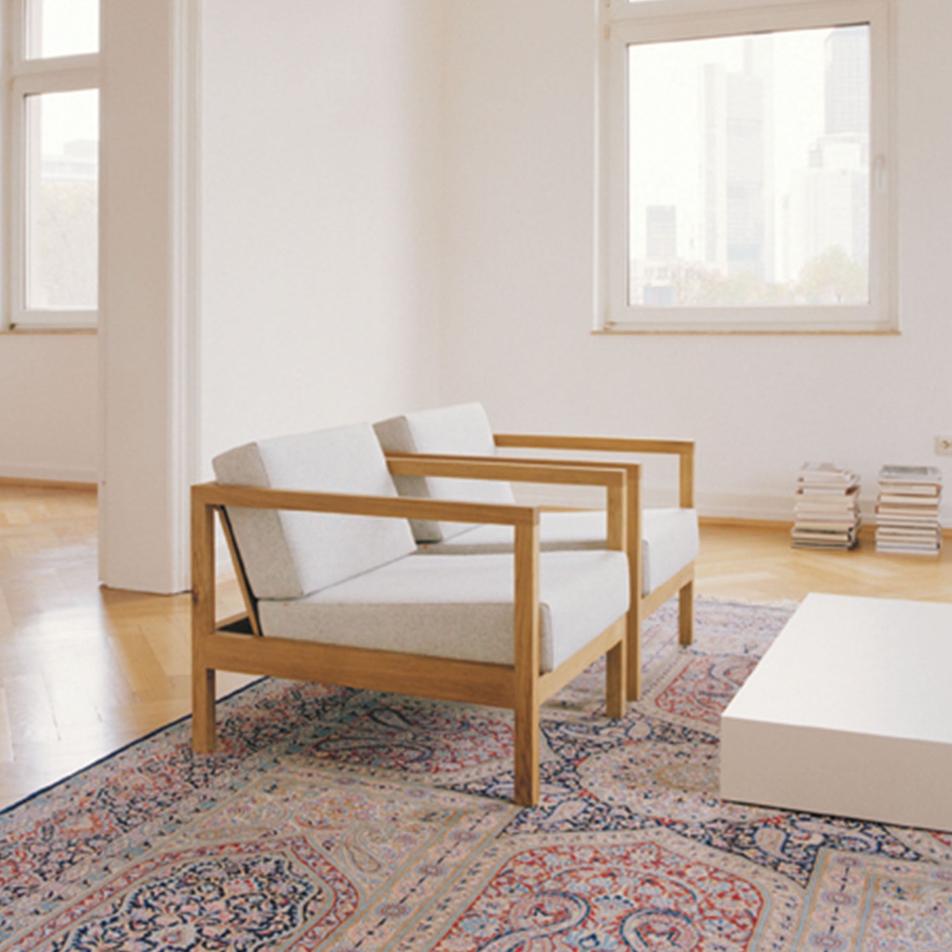 A minimalist room featuring two light gray E15 EC01 BYRON armchairs with wooden frames on a patterned rug. Large windows brighten the space, complemented by a low white table and book stack in the corner, all enhanced by natural light.